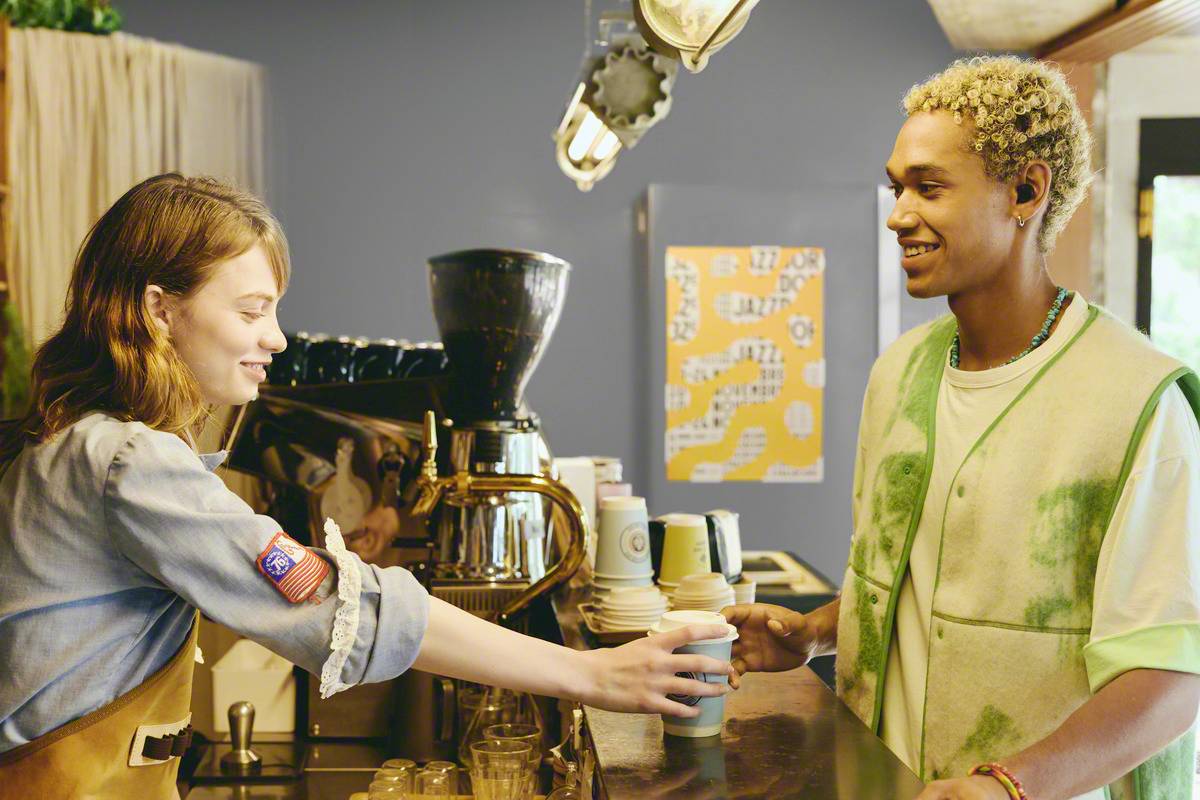 A young woman hands a young man a drink in a café. The man thanks her with a smile. In the background, a coffee machine and cups are visible.