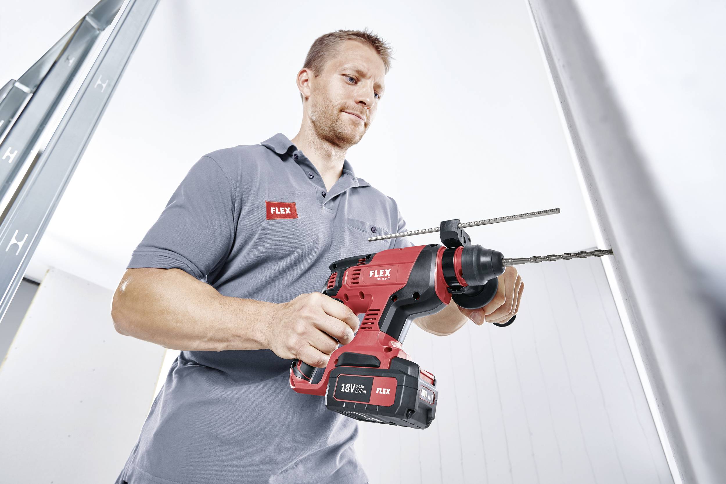 A man in a grey shirt with a 'FLEX' logo is operating a red drill, drilling into a wall. He is wearing ear protection and looks focused.