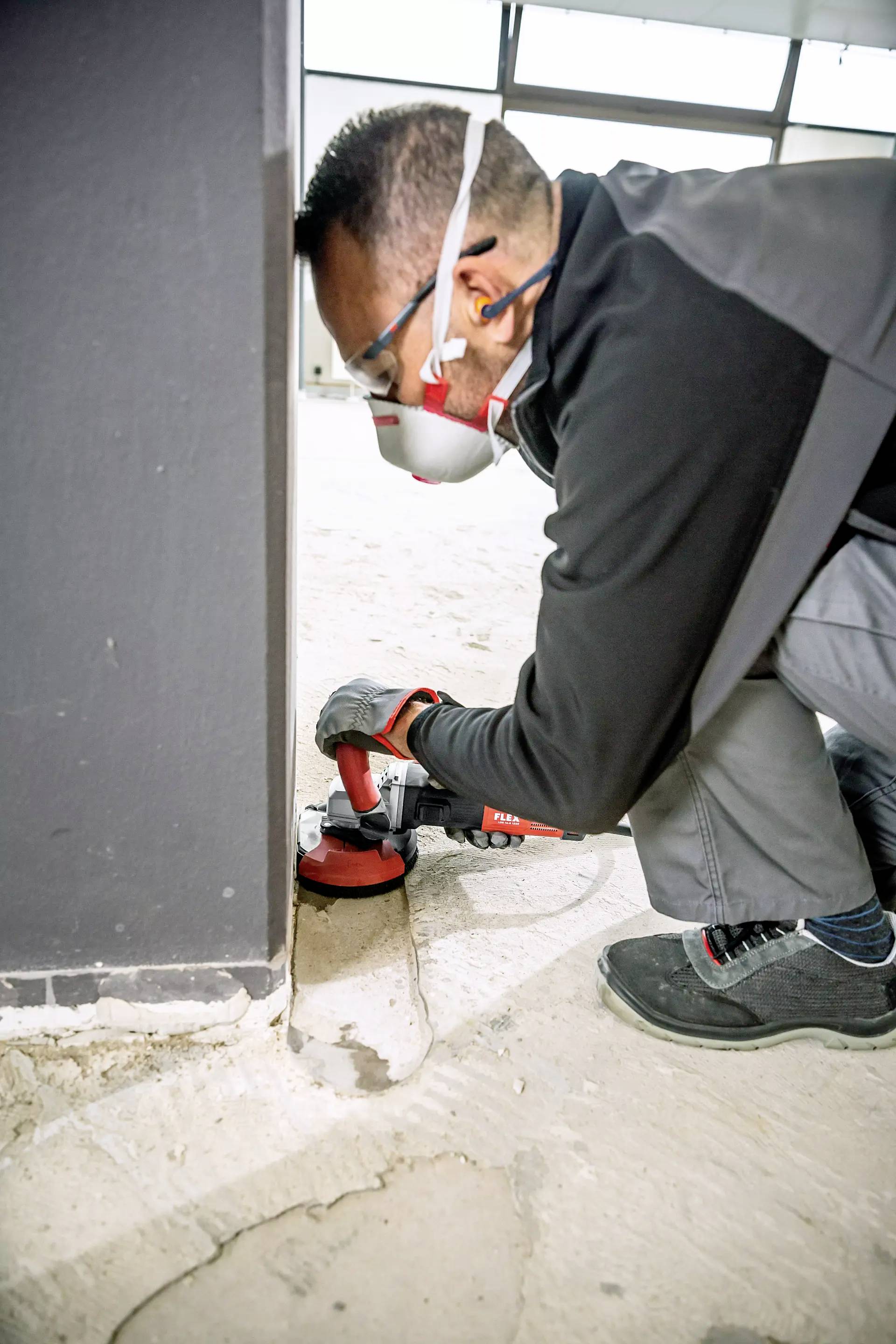 A worker wearing a protective mask and ear defenders is grinding a concrete floor with a rotating grinding machine at a building site.
