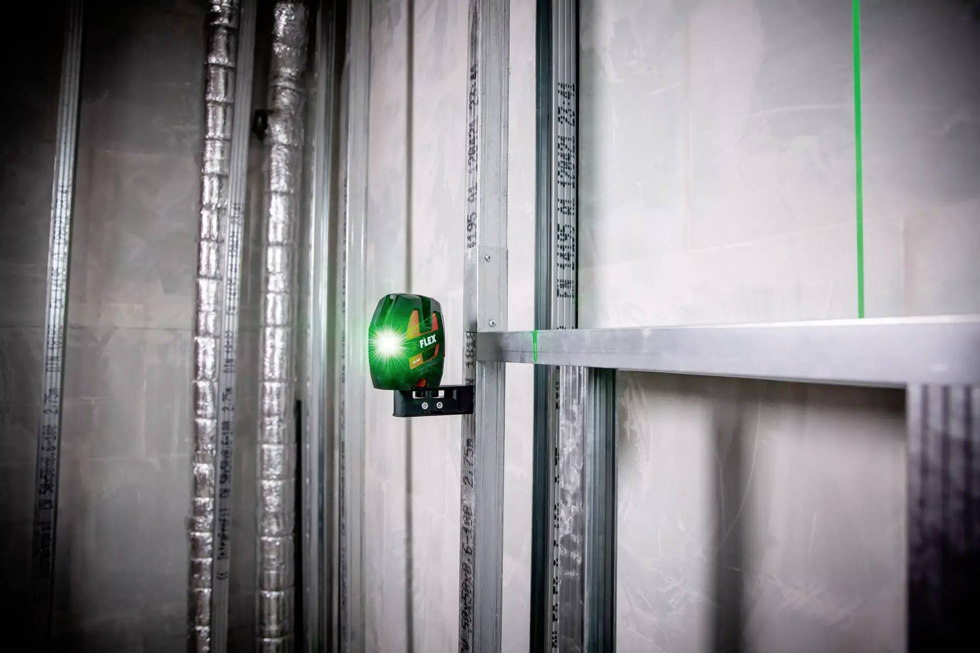 Electronic device projects green laser level onto wall in a room with metal framework and insulation to support construction work.