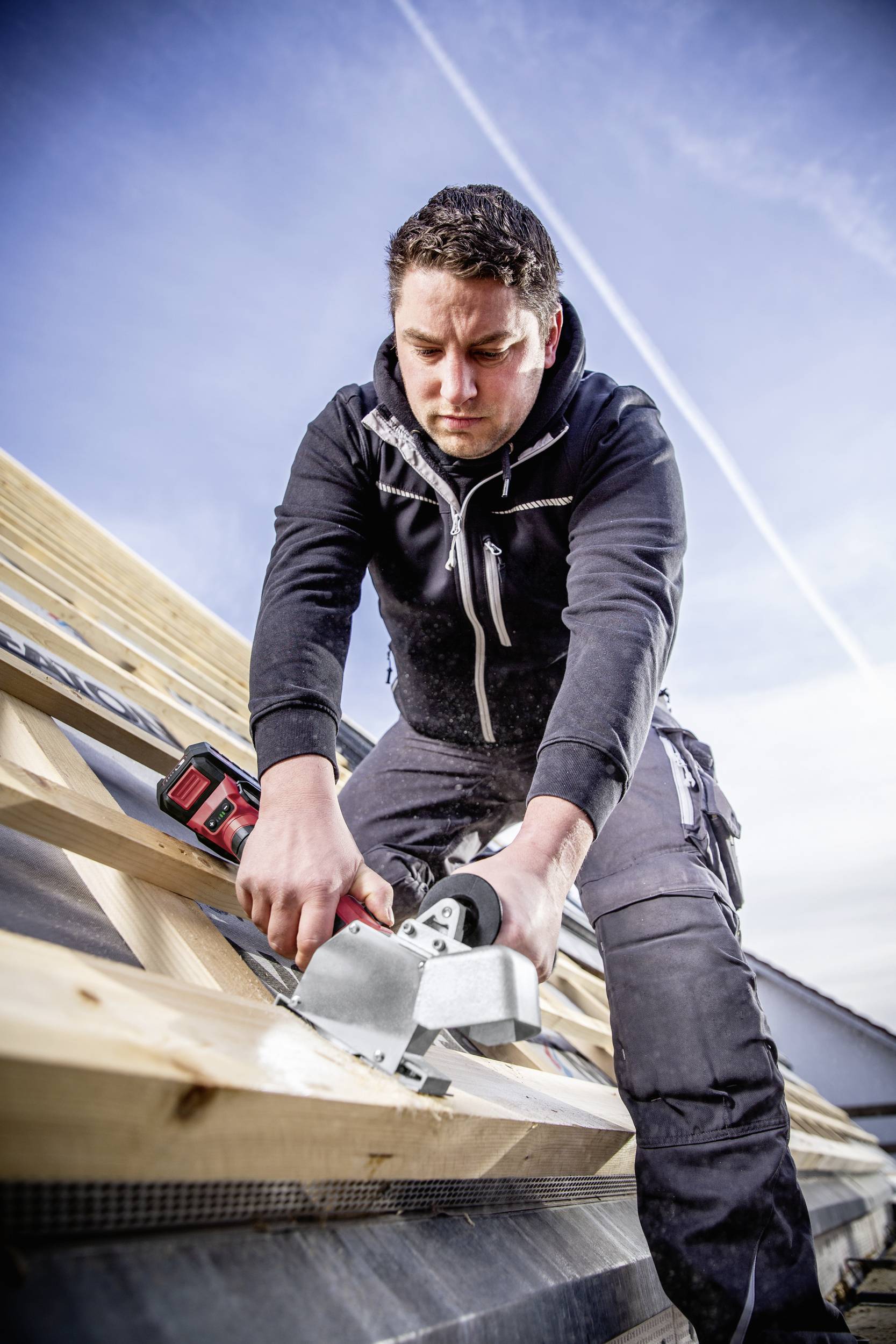 A craftsman works intently with a saw in an attic space under a clear sky, wearing dark workwear.
