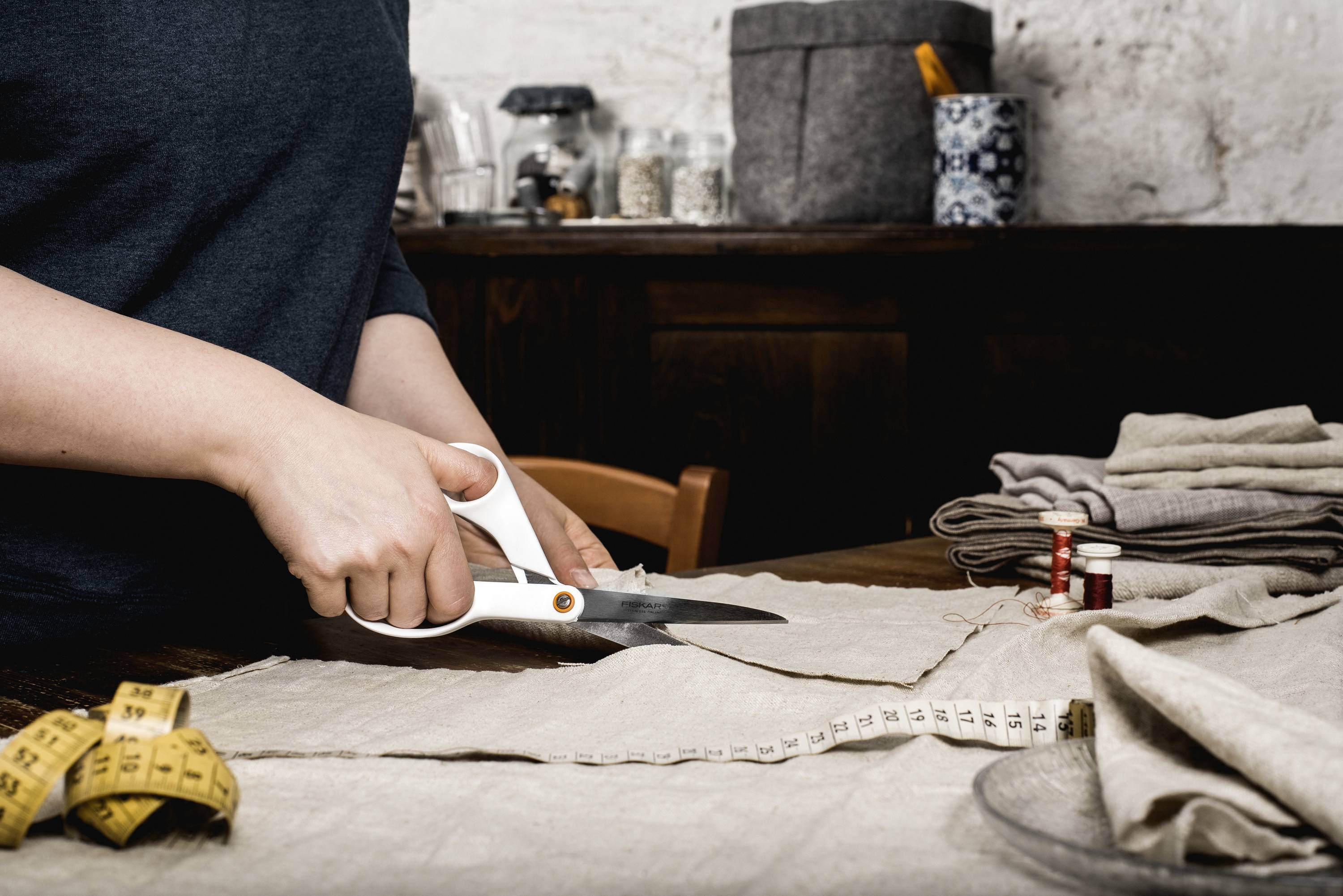 A person is cutting fabric with scissors on a table. In the background, sewing accessories such as a tape measure and thread can be seen.