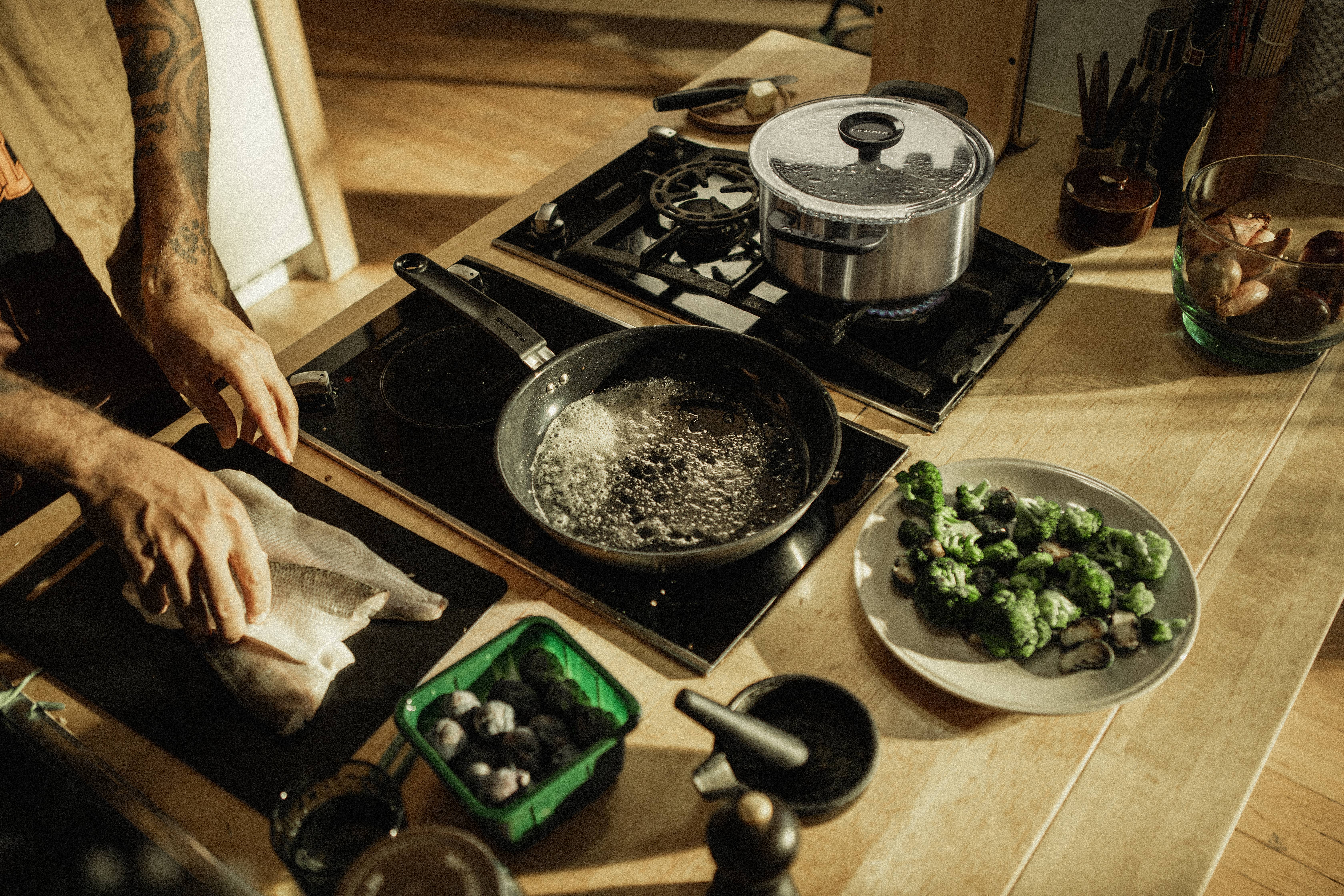 A person is cooking in a kitchen. On the hob, there is a frying pan with oil and a saucepan with a lid. On the table, there are fish fillets and Brussels sprouts.