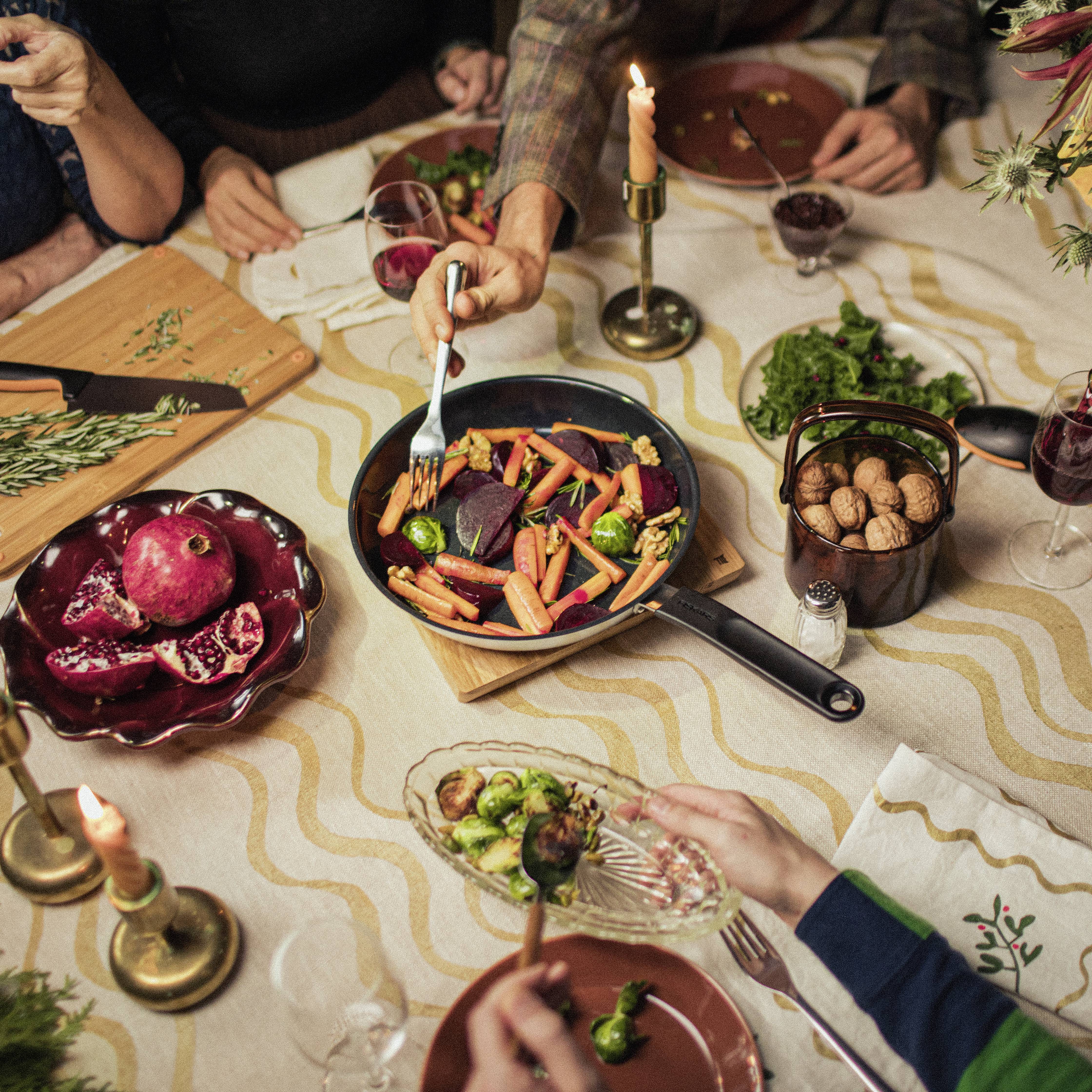 Several people are sitting around a table for dinner. A pan of colourful vegetables sits at the centre. Candles and spices are decoratively arranged.