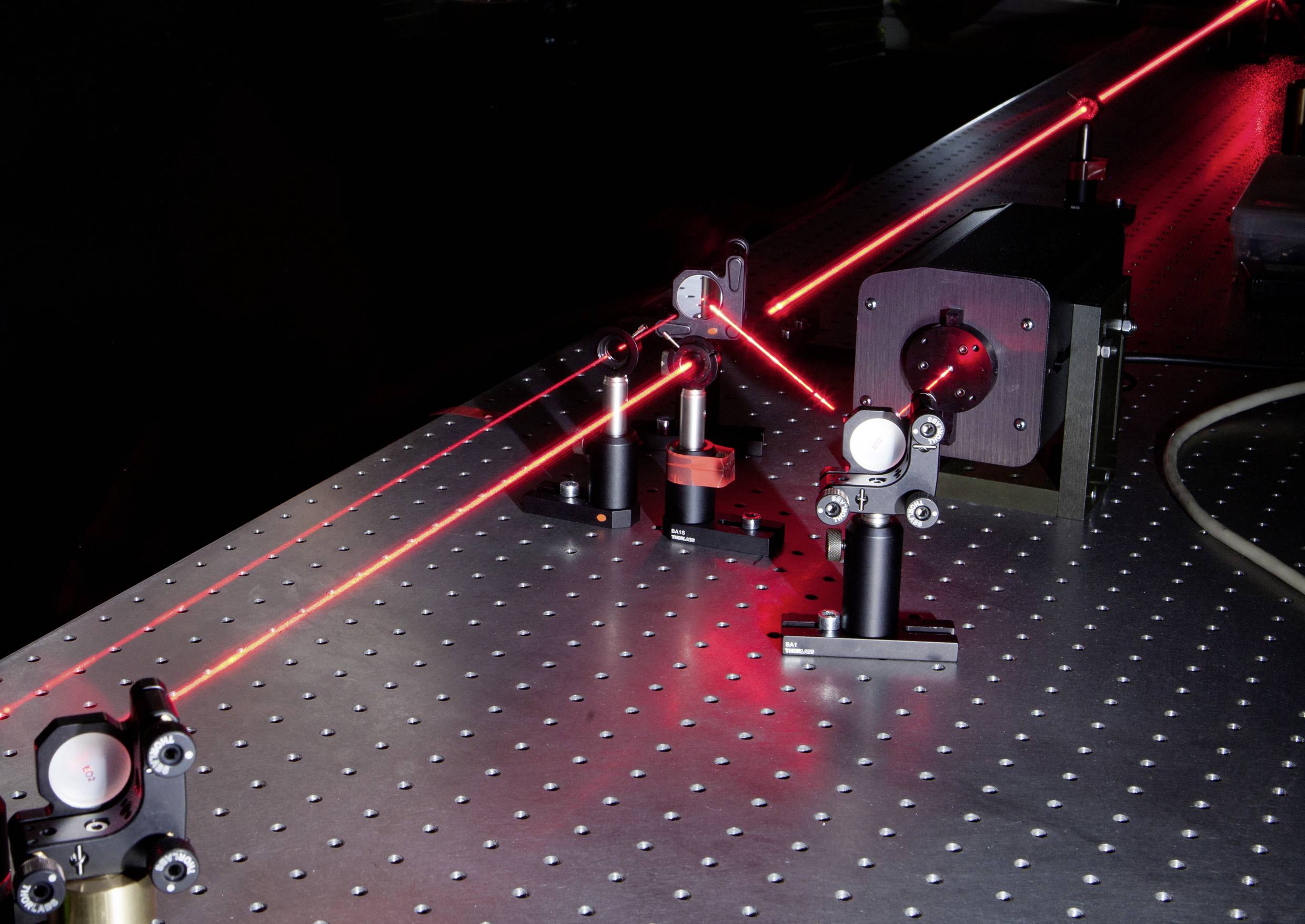 Laserstrahl trifft auf Spiegel und Linse auf einem optischen Tisch, lenkt den Strahl um. Wissenschaftliche Laborumgebung.