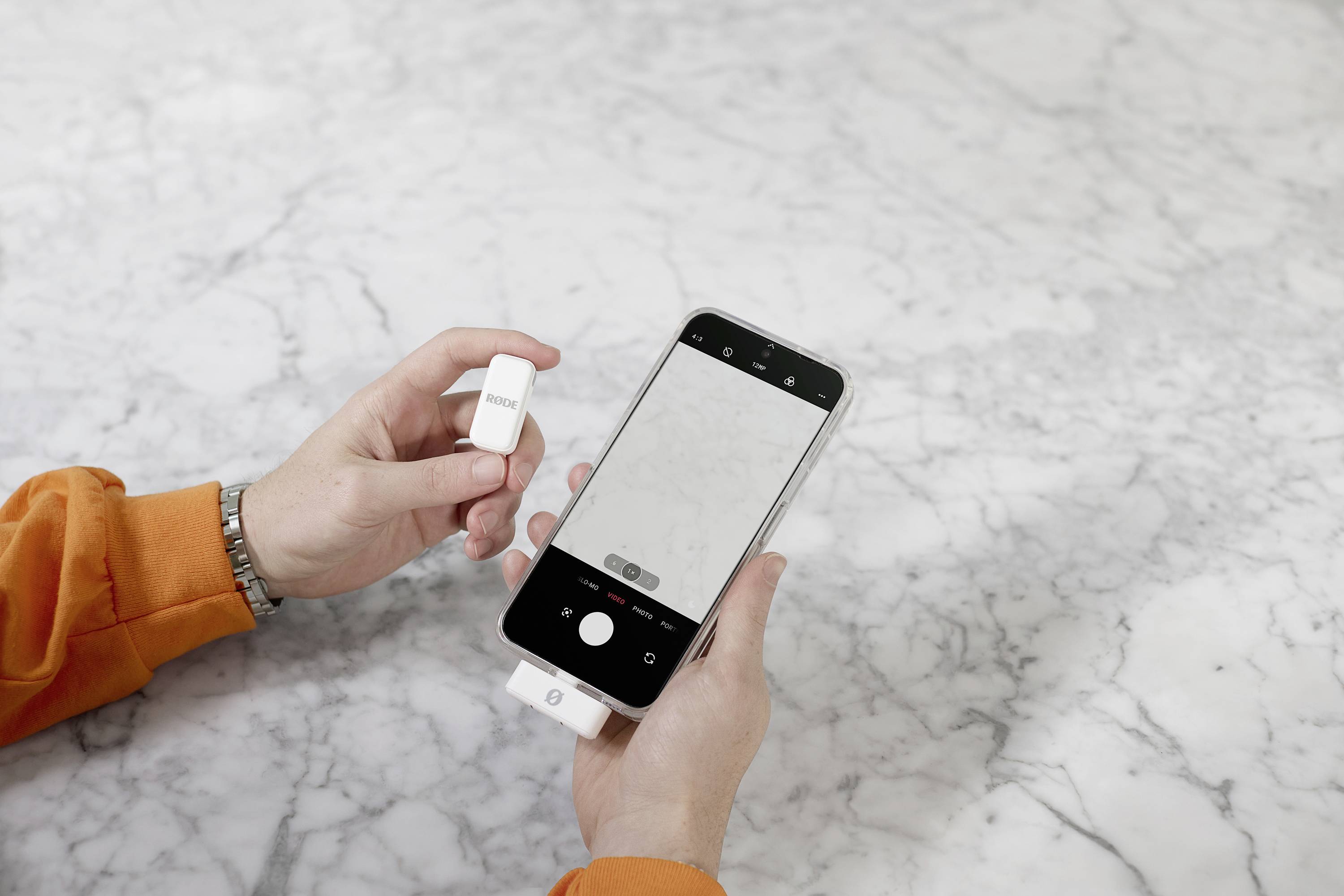 A person is holding a smartphone with an open camera app and connecting a white device to the mobile phone. A marble background is in focus.
