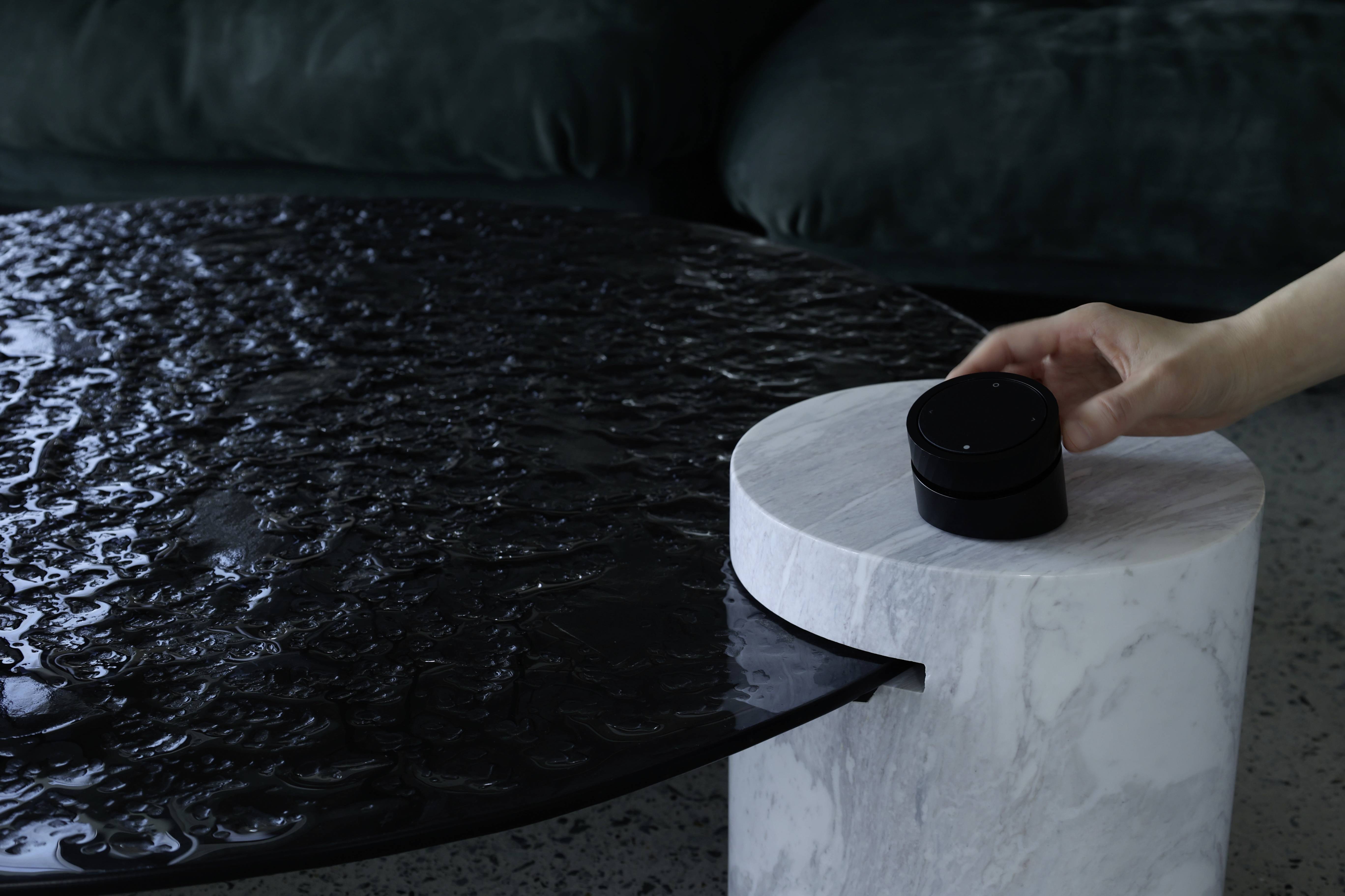 A hand reaches for a black round object on a white, marble-like plinth beside a dark table surface.