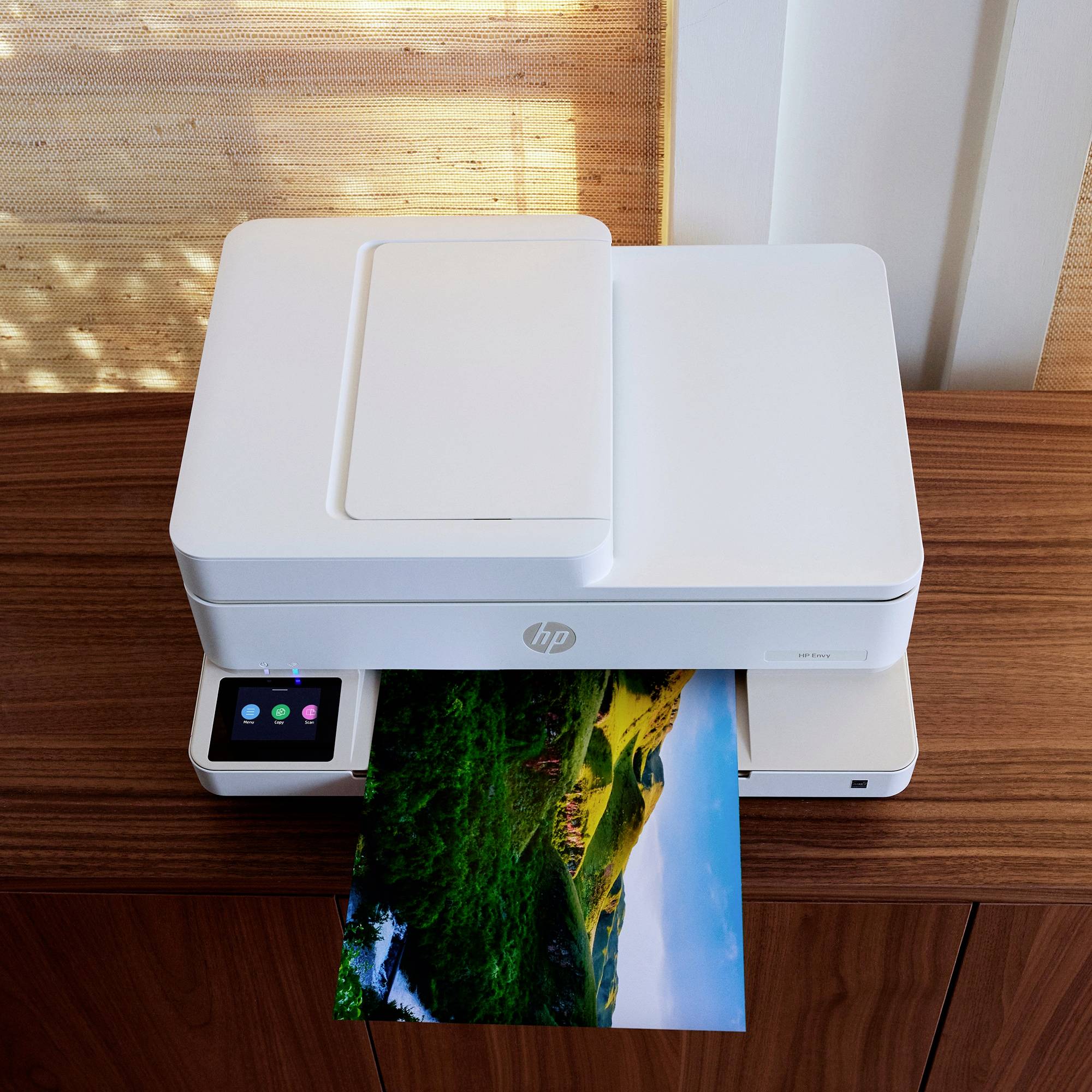A white printer on a wooden table is printing a photo with a landscape in vibrant green tones. On the left is a control panel.