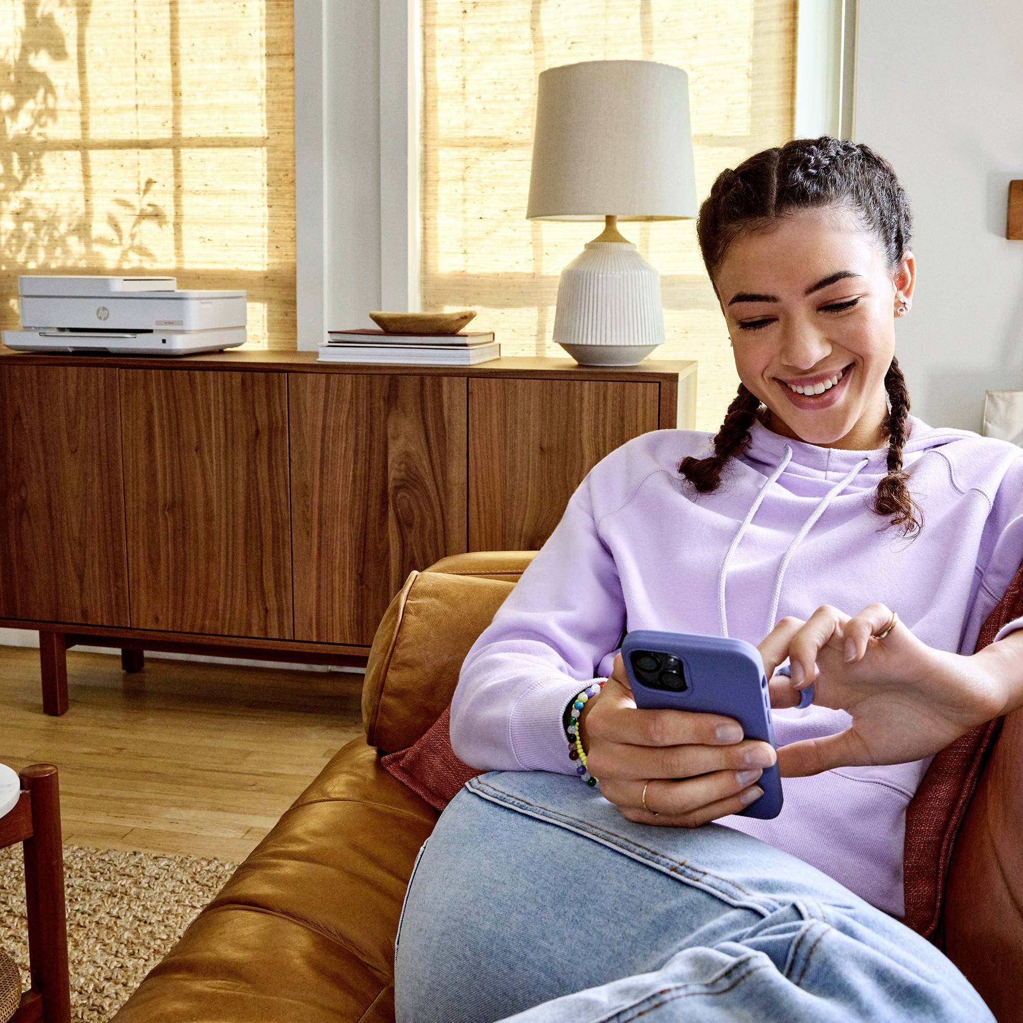 A smiling person is sitting on a sofa and using a smartphone, with a printer and plants in the background of a living room.