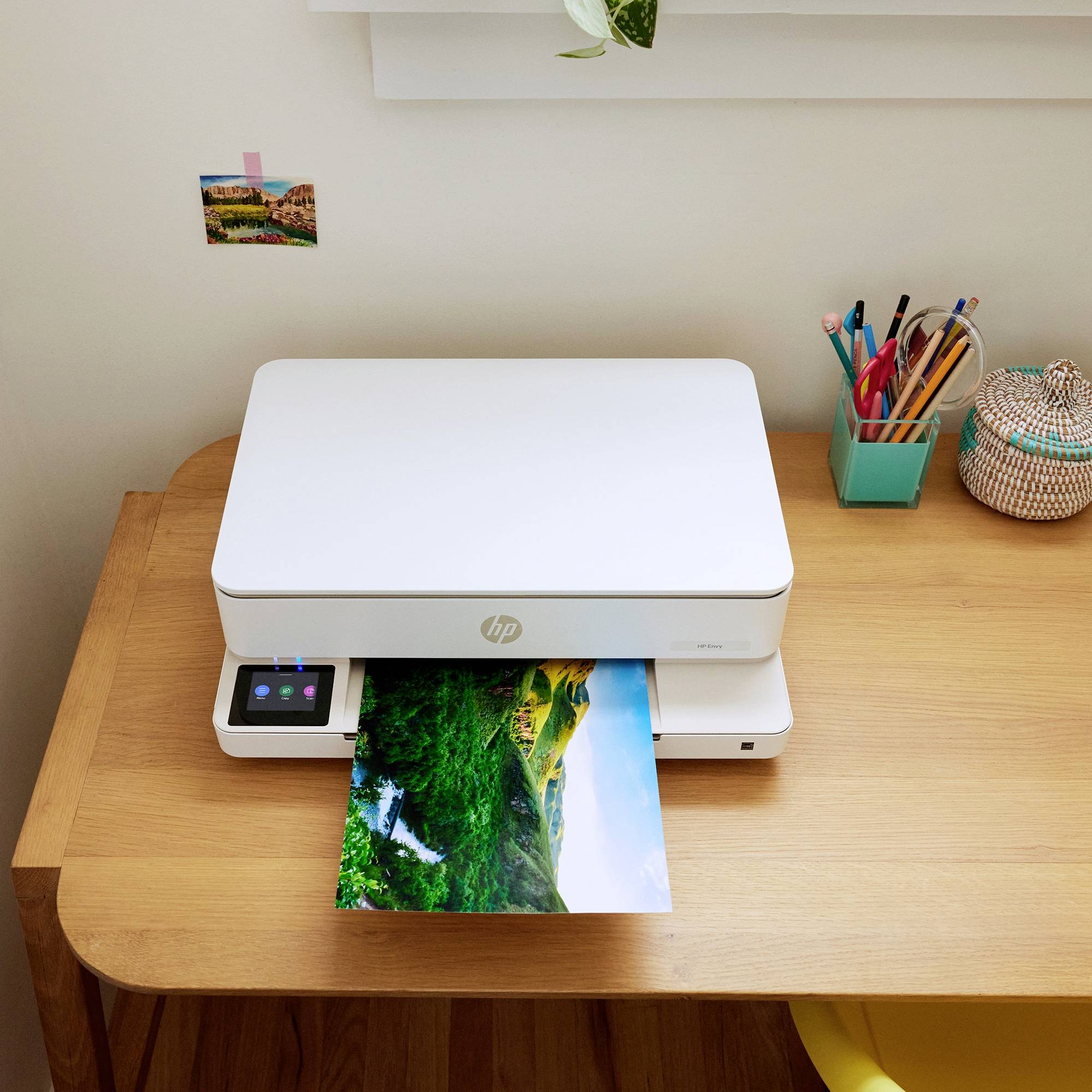 A printer sits on a desk and is printing a photo of a landscape scene. Next to the printer, pens are placed in a container.