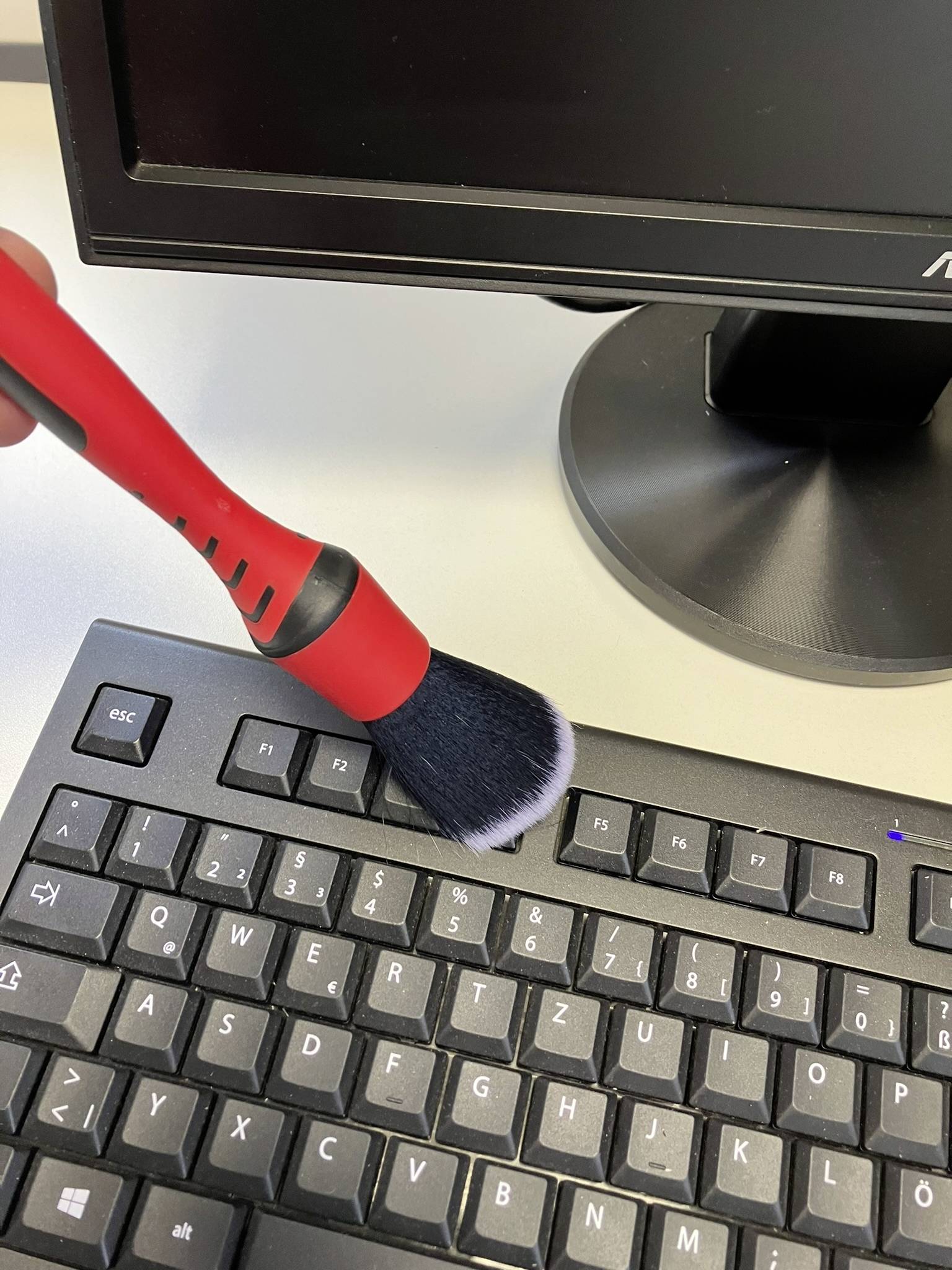 A hand uses a red and black brush to remove dust from a computer keyboard. A monitor is visible in the background.