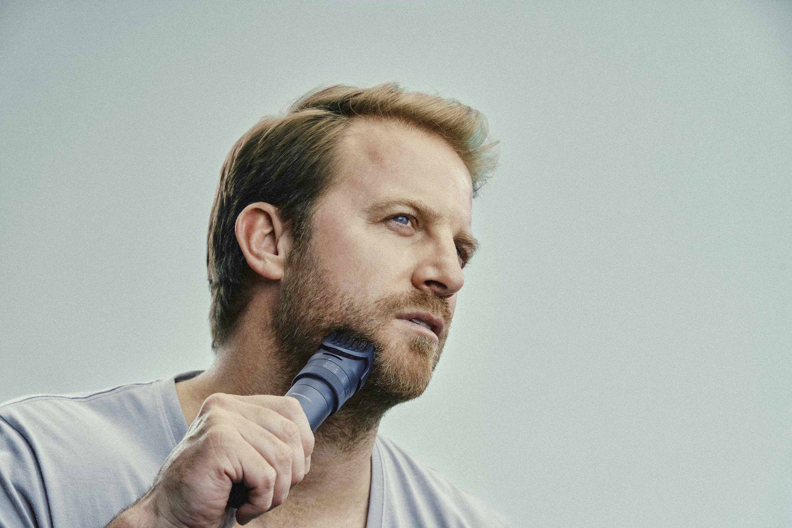 A person with brown hair is shaving their beard with an electric razor and looking to the side.