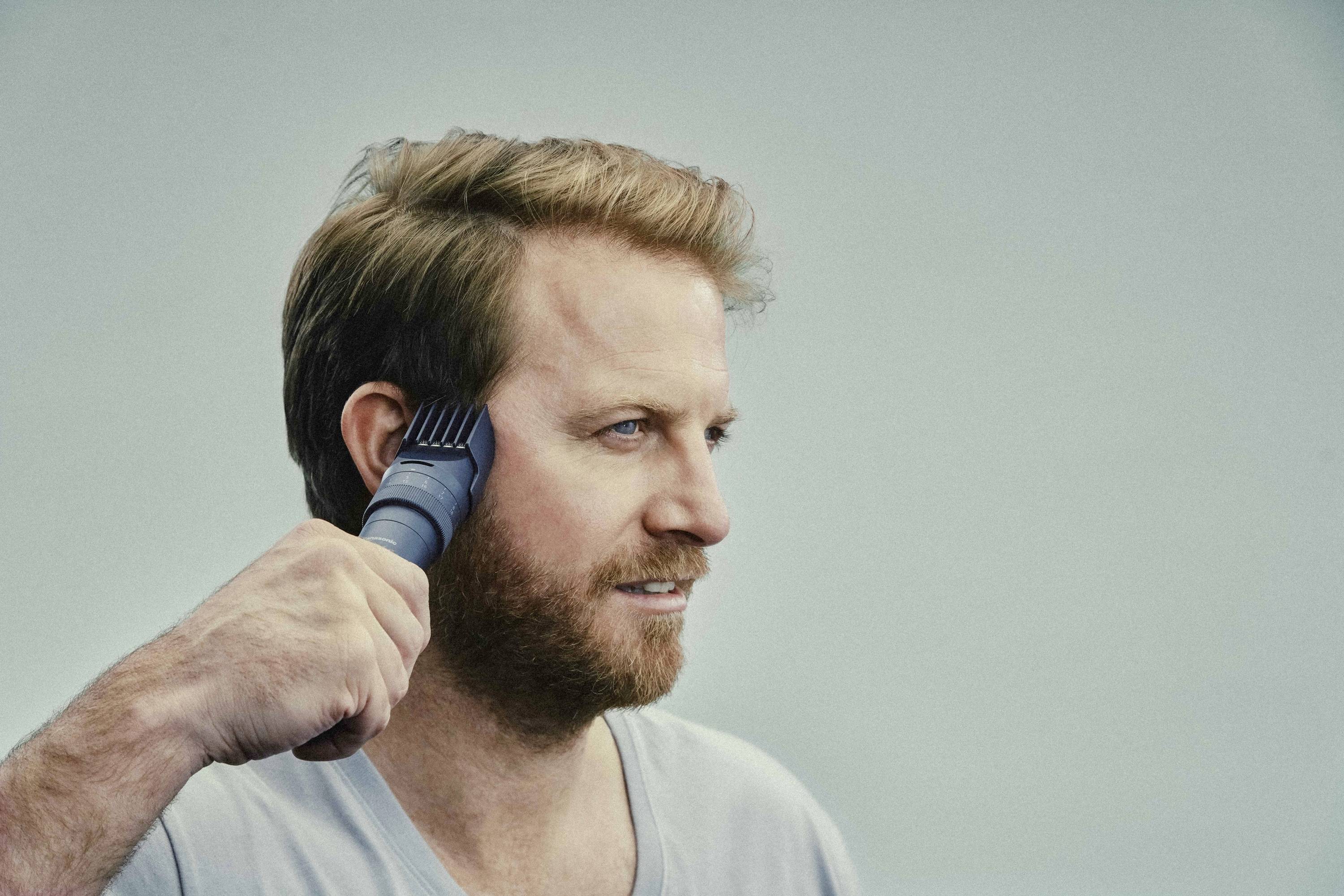 A man is cutting his hair with an electric hair trimmer. He has short, brown hair and is wearing a grey T-shirt.