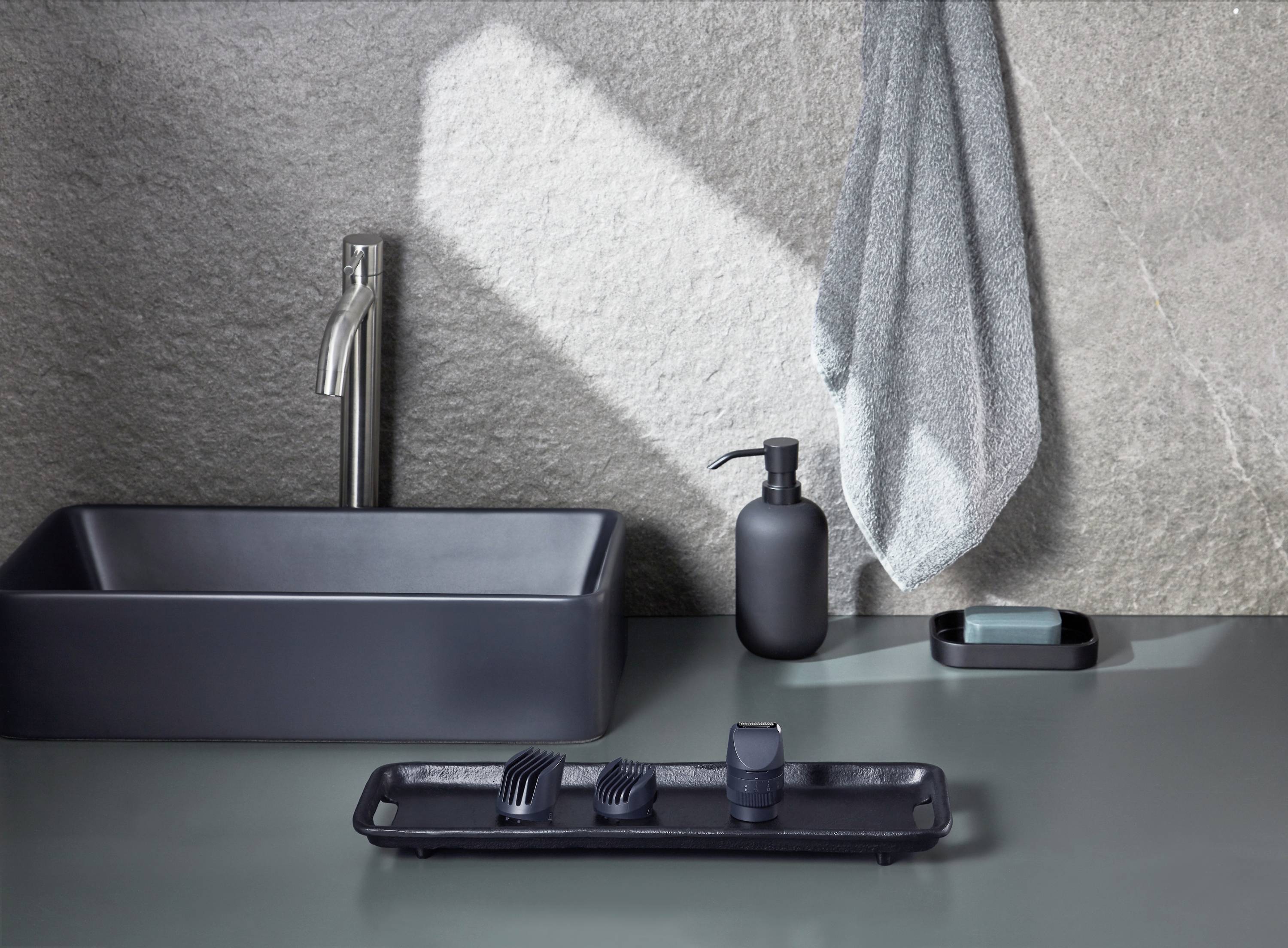 Washbasin scene with a dark, angular washbasin, soap dispenser, soap dish, and folded towels standing on a modern worktop.