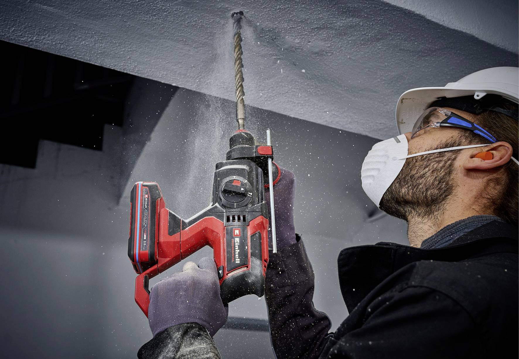 A person in protective clothing is drilling a hole into a concrete beam with a red cordless drill. Dust is spraying from the drill hole.