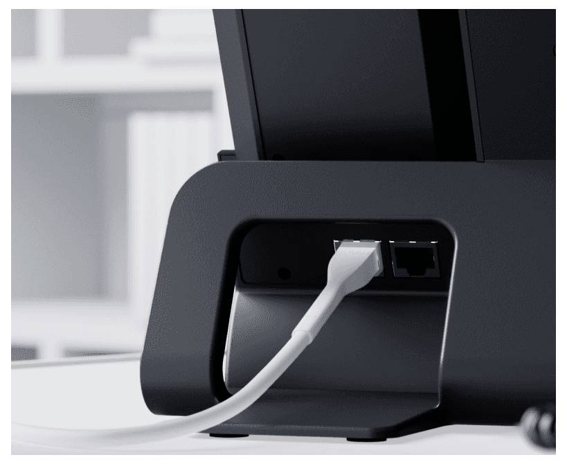 An Ethernet cable is connected to the rear port of a monitor, with a blurred bookshelf in the background.