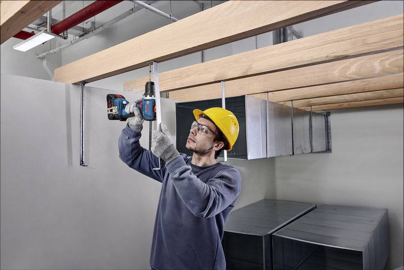 A construction worker wearing a hard hat is securing metal braces to a ceiling using a cordless drill during a building construction project.