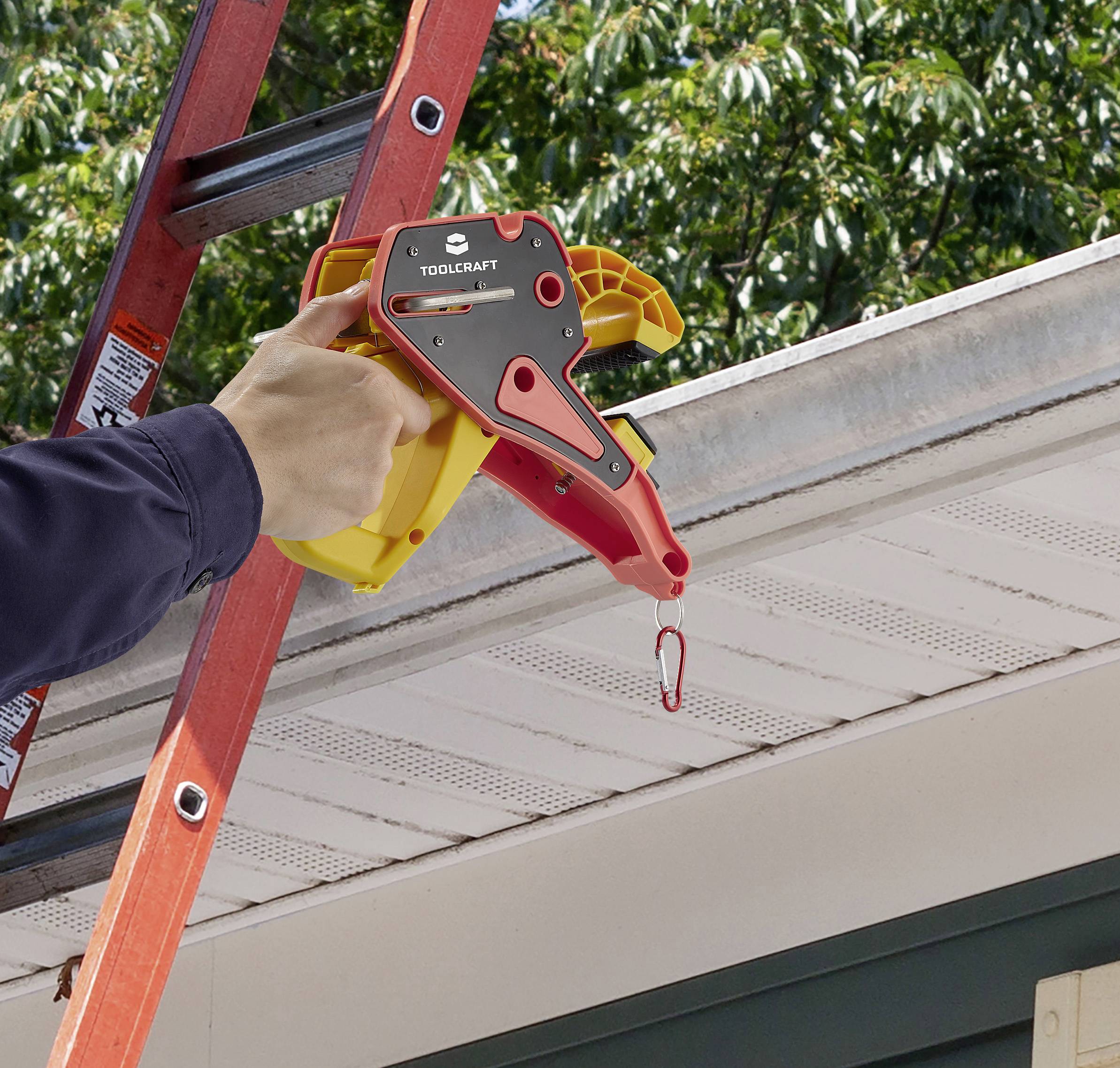 A person is attaching a red and yellow gutter cleaner to a guttering whilst standing on a ladder. Trees can be seen in the background.
