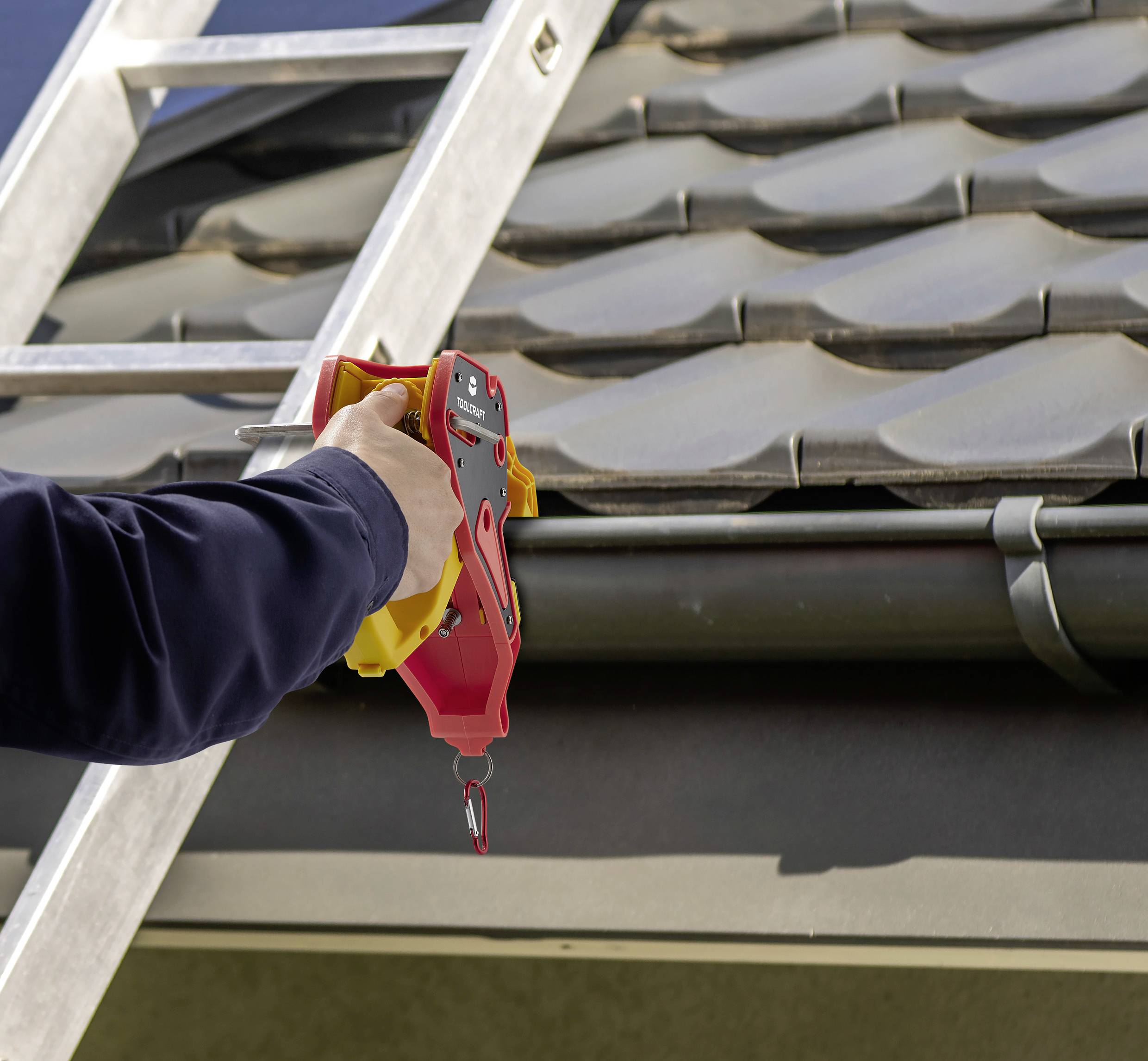 A person is securing a safety device to a gutter, next to a ladder leaning against the roof edge, in front of a tiled roof.