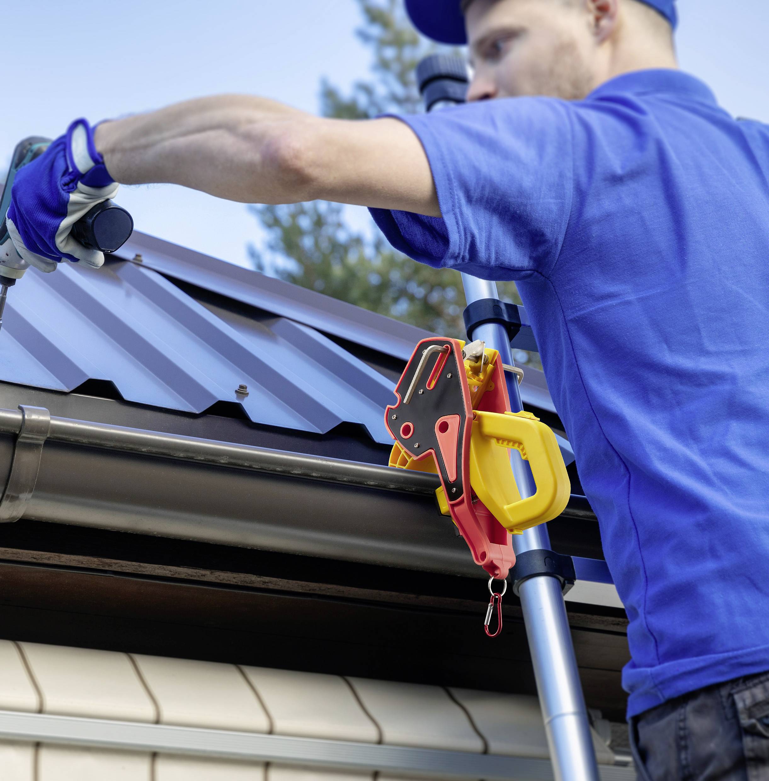 A worker in a blue uniform is using tools on a metal roof. Safety equipment is visible, with trees in the background.