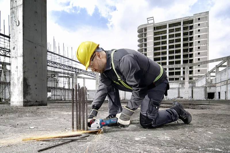 A construction worker in safety gear cuts metal rods with a power tool at a building site, with a partially constructed high-rise in the background.
