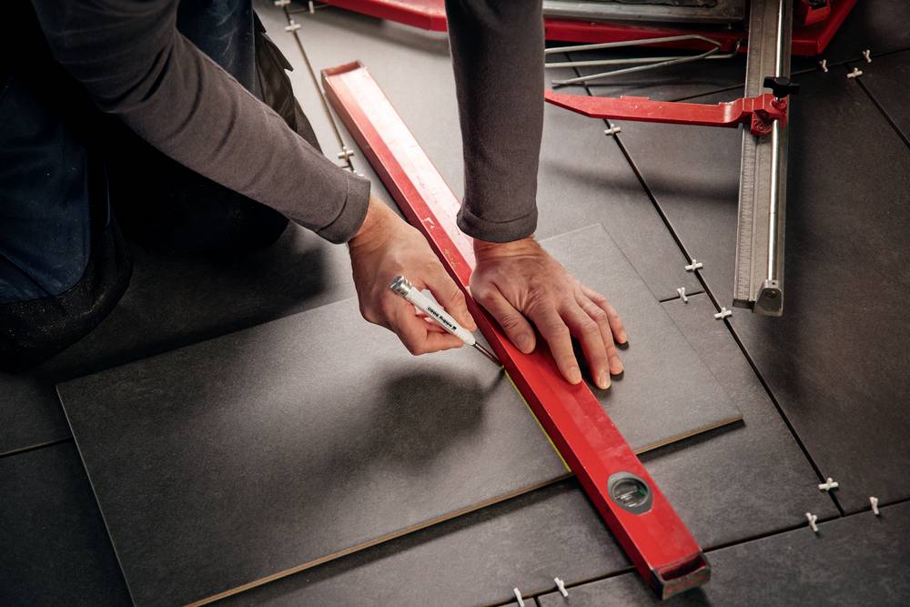 A person is precisely cutting a ceramic tile using a red ruler on a tiled floor.