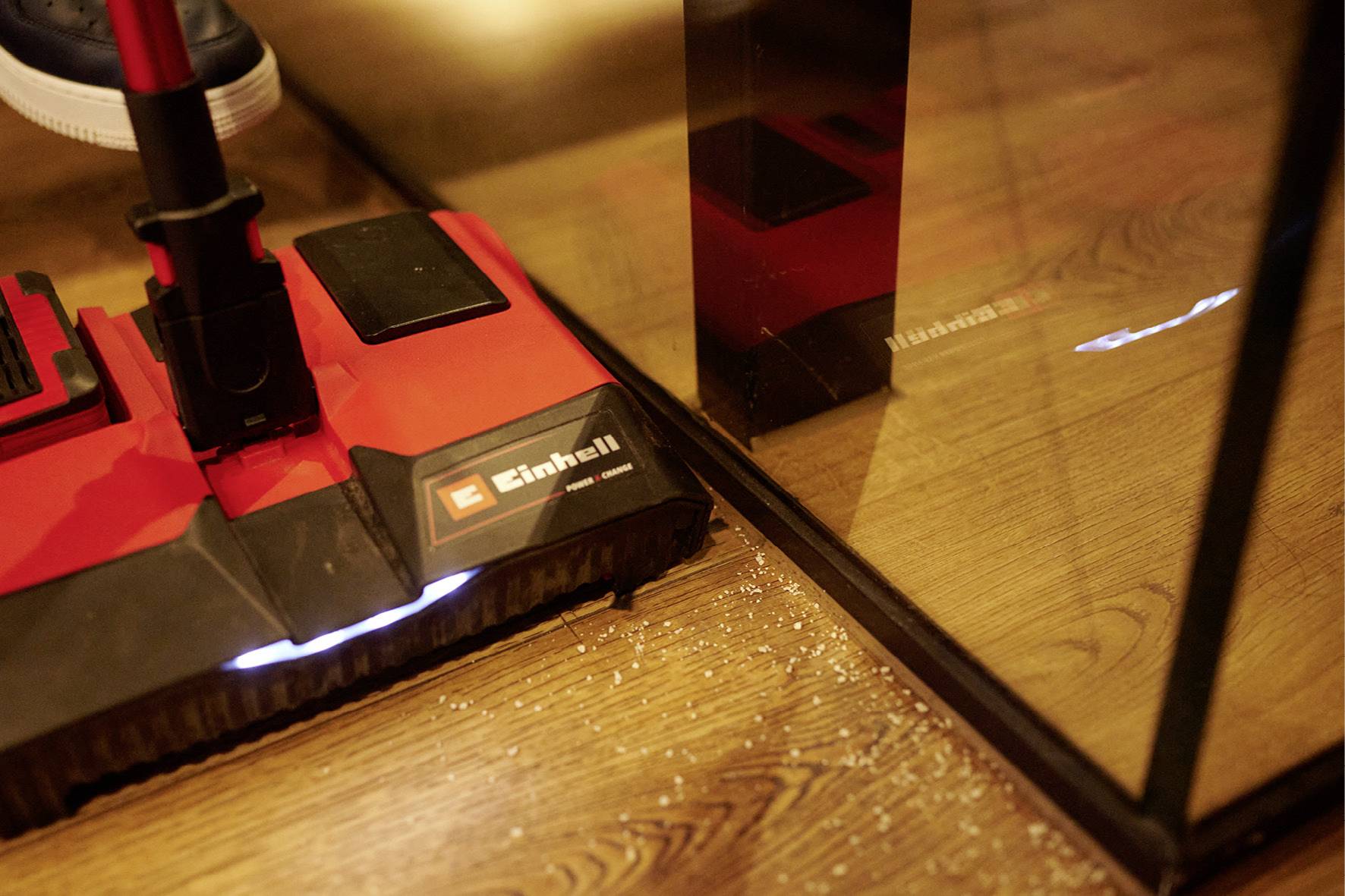 A red Einhell vacuum cleaner is sucking up a wooden floor and removing small white particles. A shoe is visible in the top left corner.