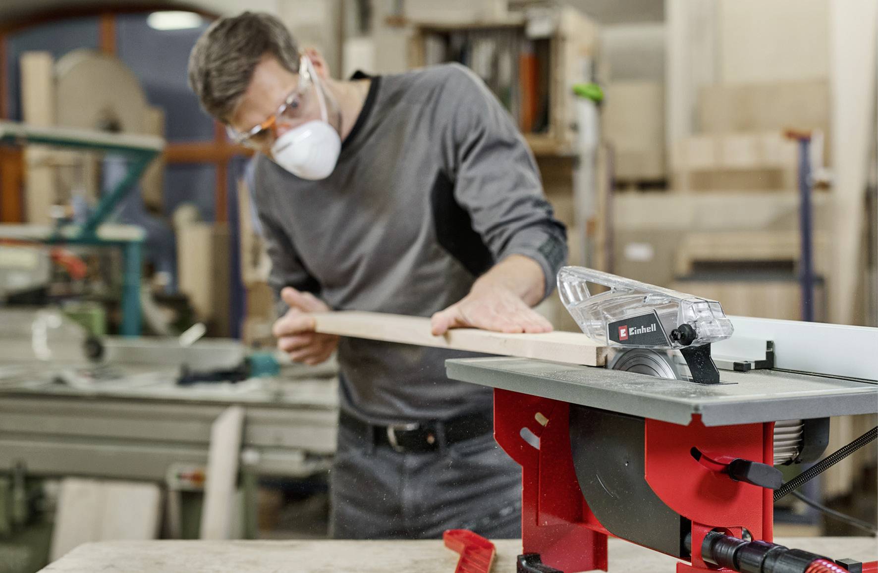 A man wearing safety glasses and a mask is working on a wooden board in a carpenter's workshop. A circular saw is in the foreground.