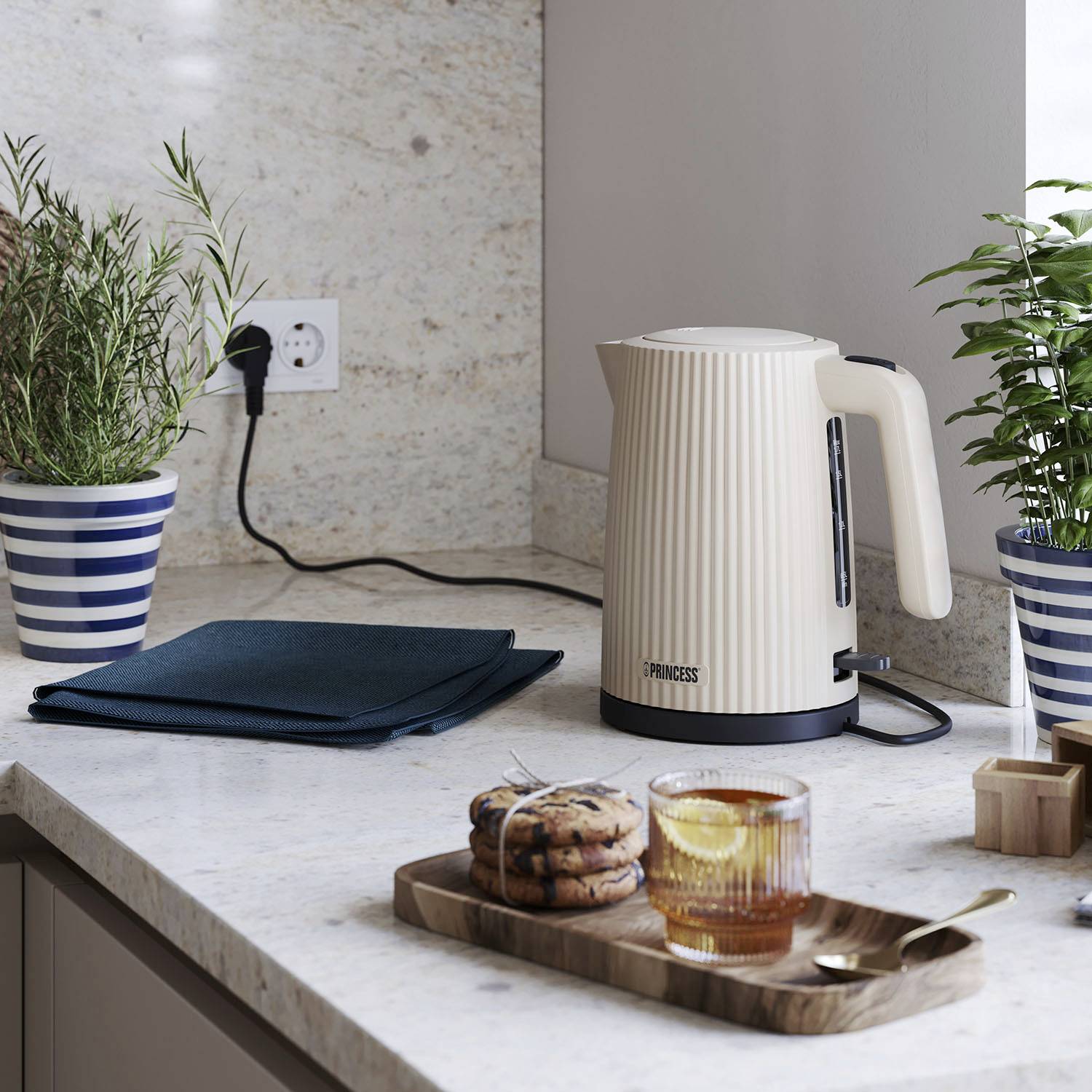 A kettle is sitting on a kitchen worktop next to plants and a mug with a drink and biscuits on a wooden board.
