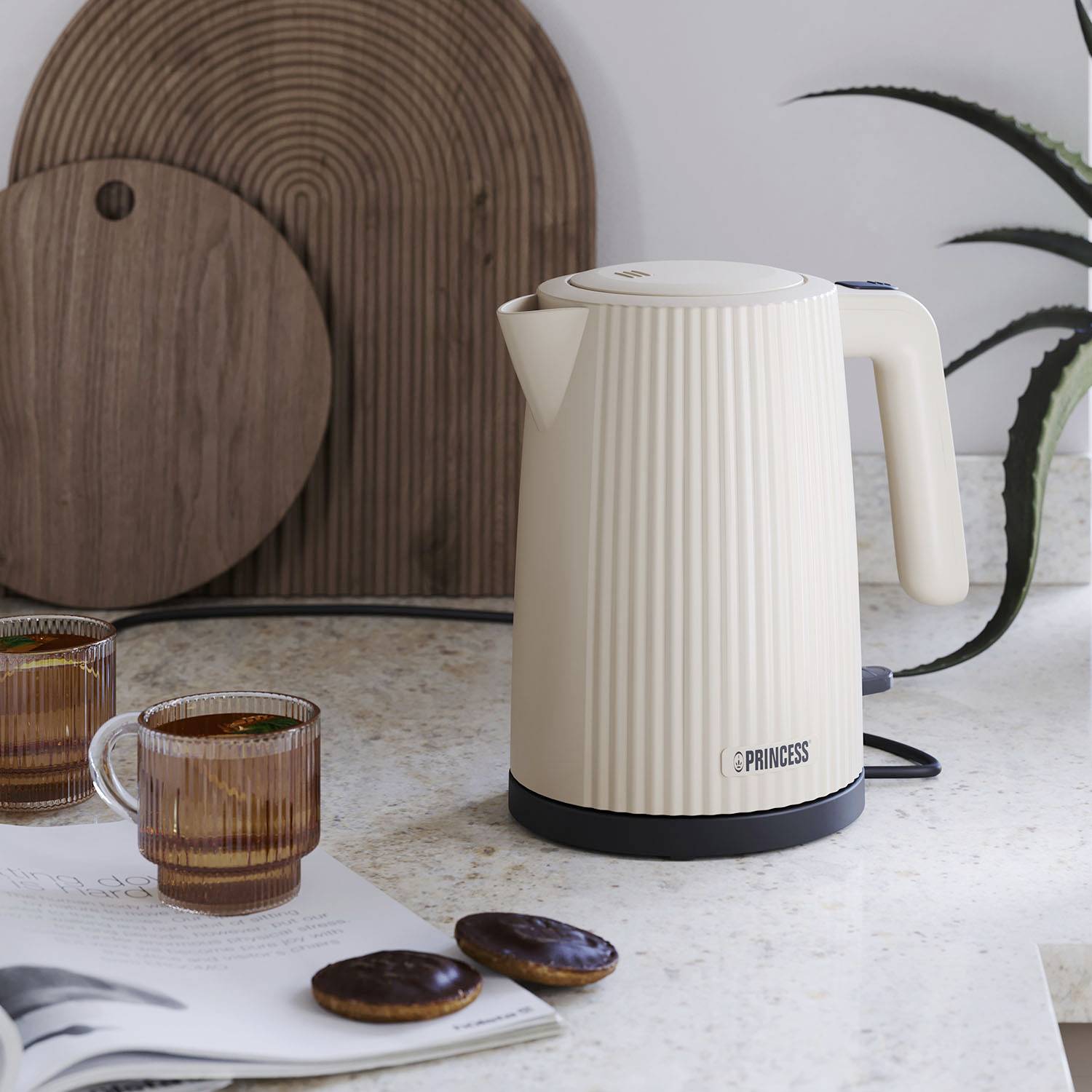 A beige kettle on a kitchen worktop, surrounded by two glasses and two biscuits. In the background is a wooden board and a plant.