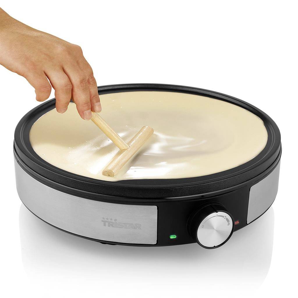 A hand spreads liquid batter on a round, heated crêpe pan using a wooden spatula.