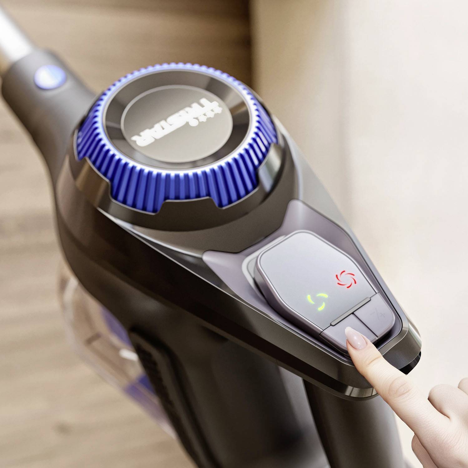 A hand presses the power button of a cordless vacuum cleaner on a wooden floor. The vacuum cleaner has blue and black details.