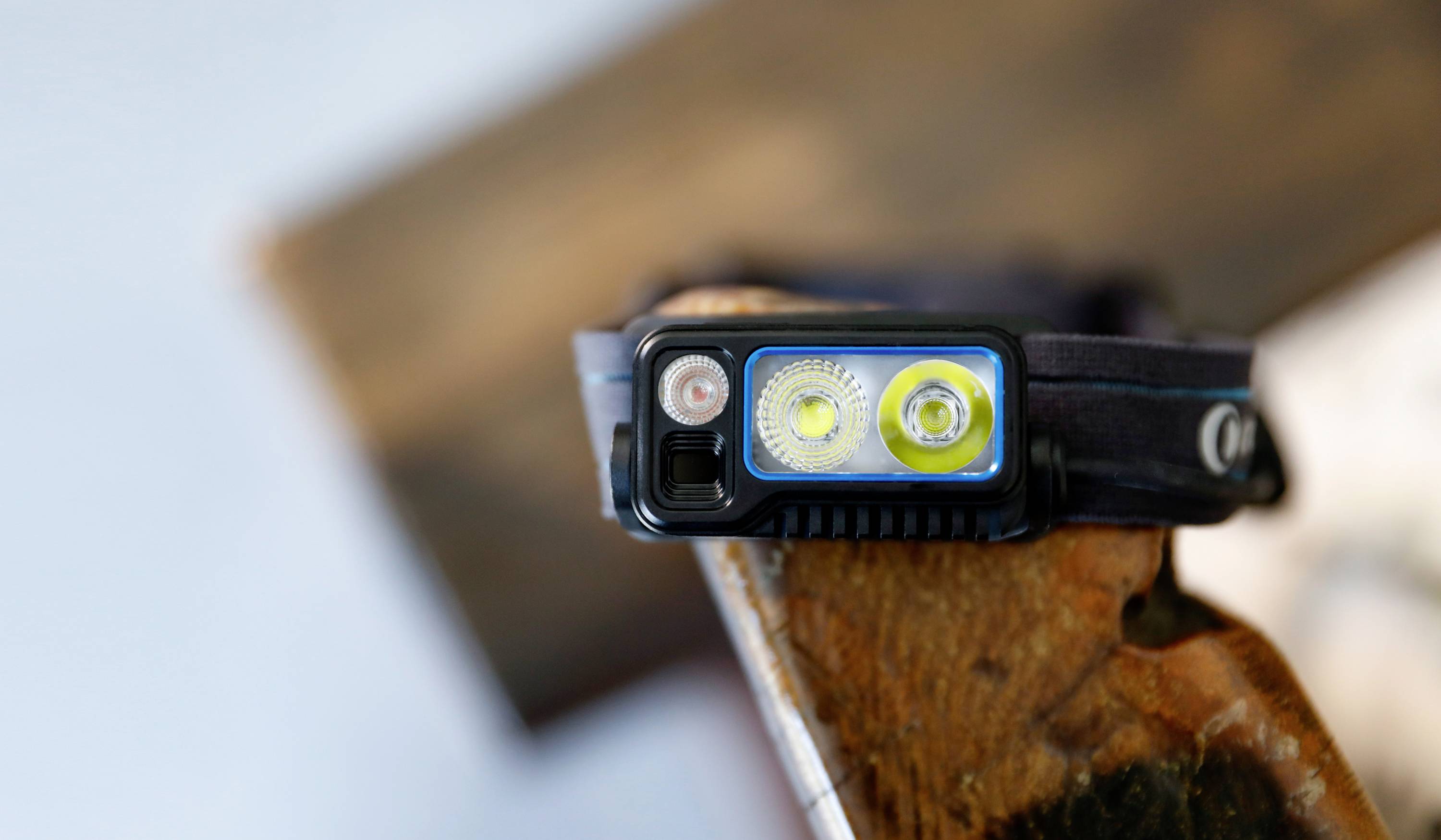 A black head torch with three LED light sources is placed on a piece of wood. It appears shock-resistant and robust for outdoor activities.