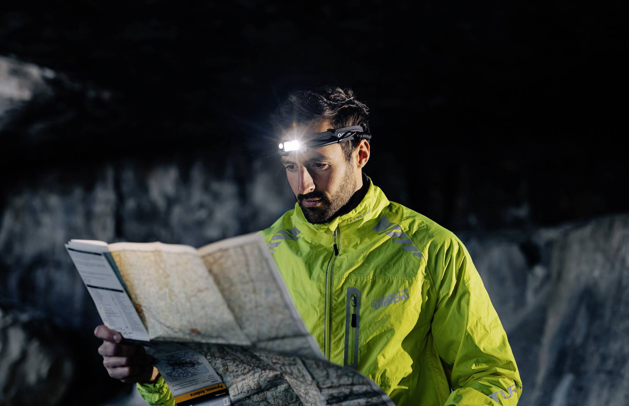 A man in a high-visibility jacket is reading a map with a head torch in a dark cave.