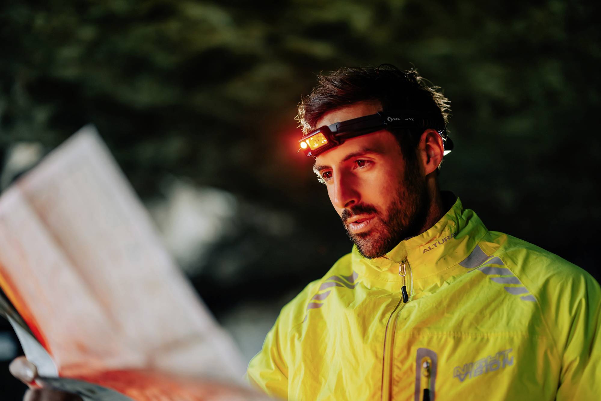 A man wearing a yellow jacket studies a map intently, wearing a head torch in a dark environment. Adventure or exploration seems possible.