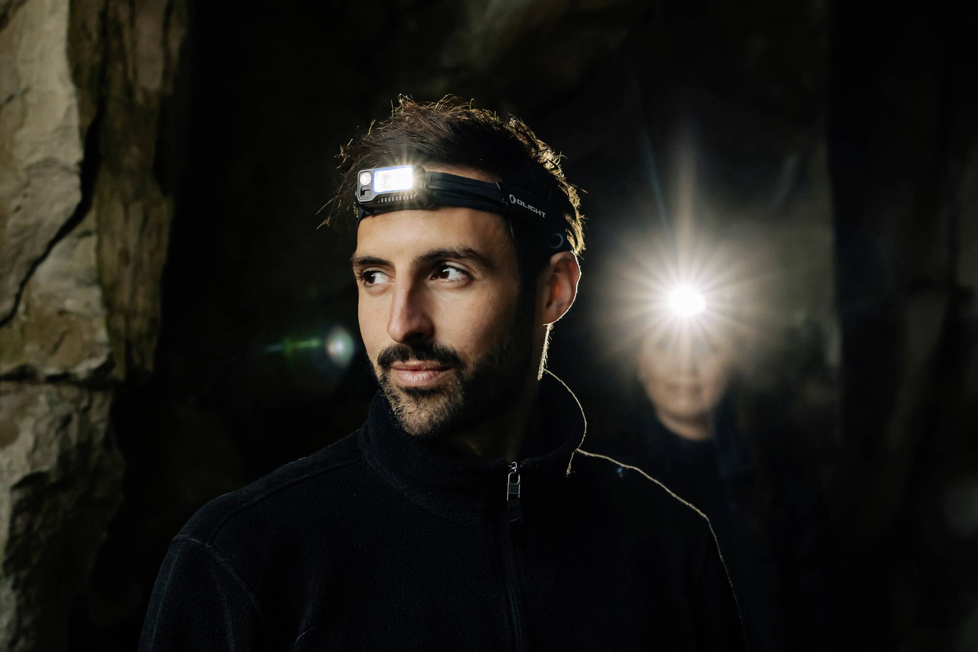 A man wearing a head torch stands in a cave, with another person in the background also wearing a head torch.