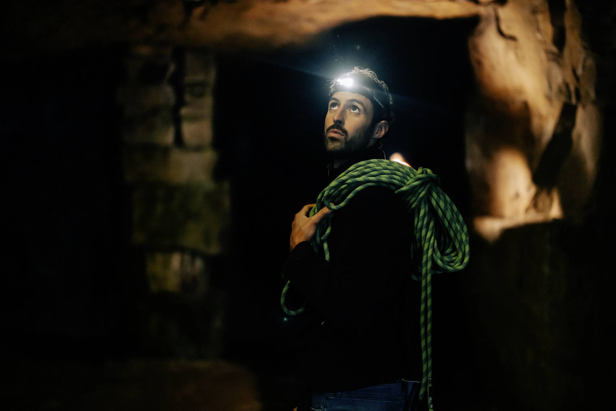 A man wearing a head torch and carrying a climbing rope looks upwards inside a cave, illuminated by warm light. Dark and rustic surroundings.