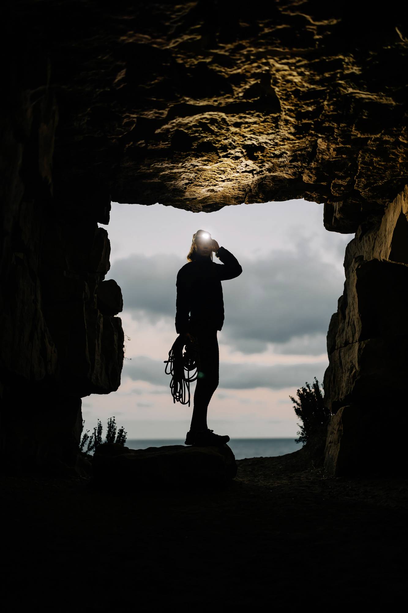 A person wearing a head torch stands in a cave, gazing out towards the sea at sunset, holding a climbing rope in their hand.