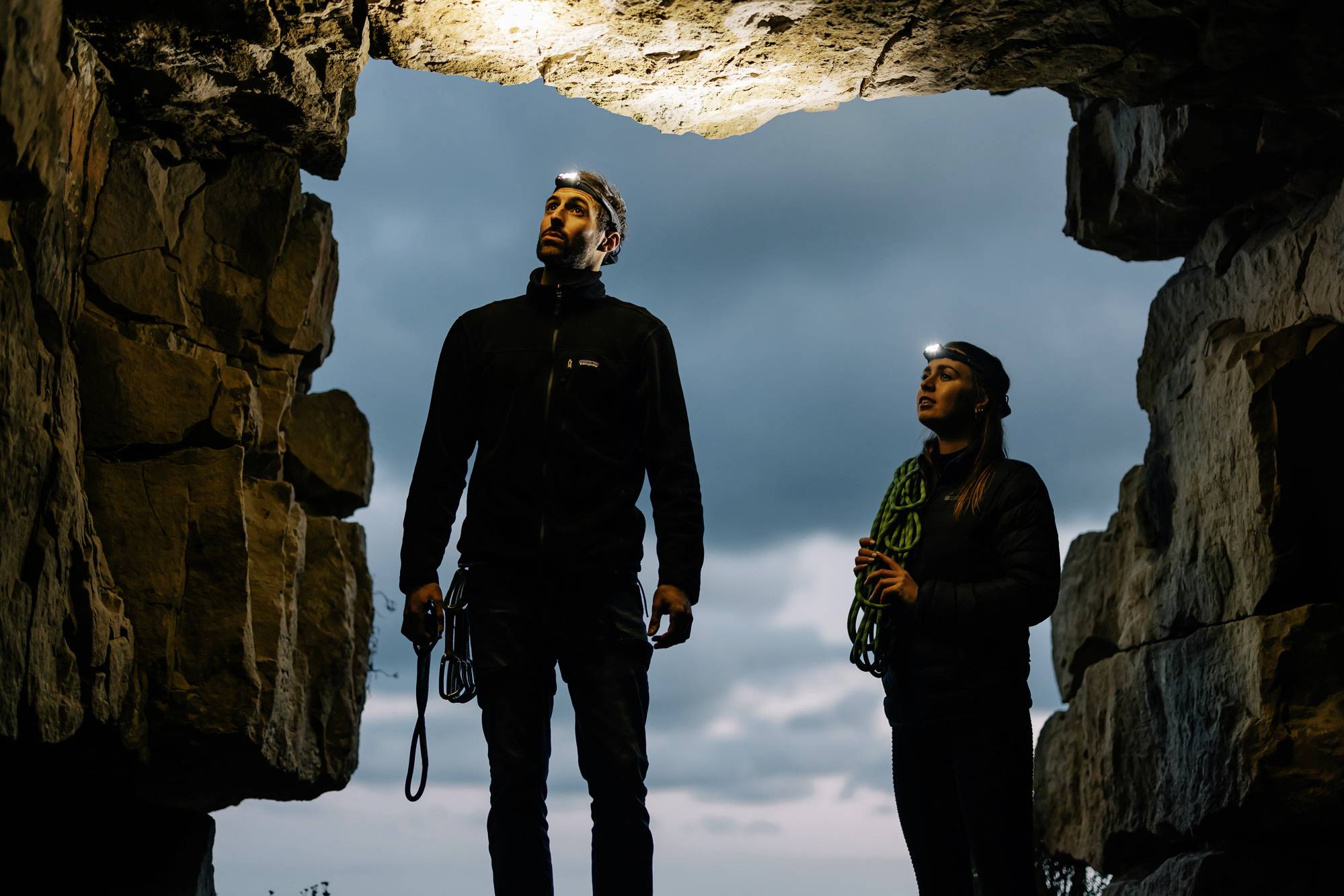Two people with head torches stand in a rocky opening outdoors. They look upwards, surrounded by rocks, beneath an overcast sky.