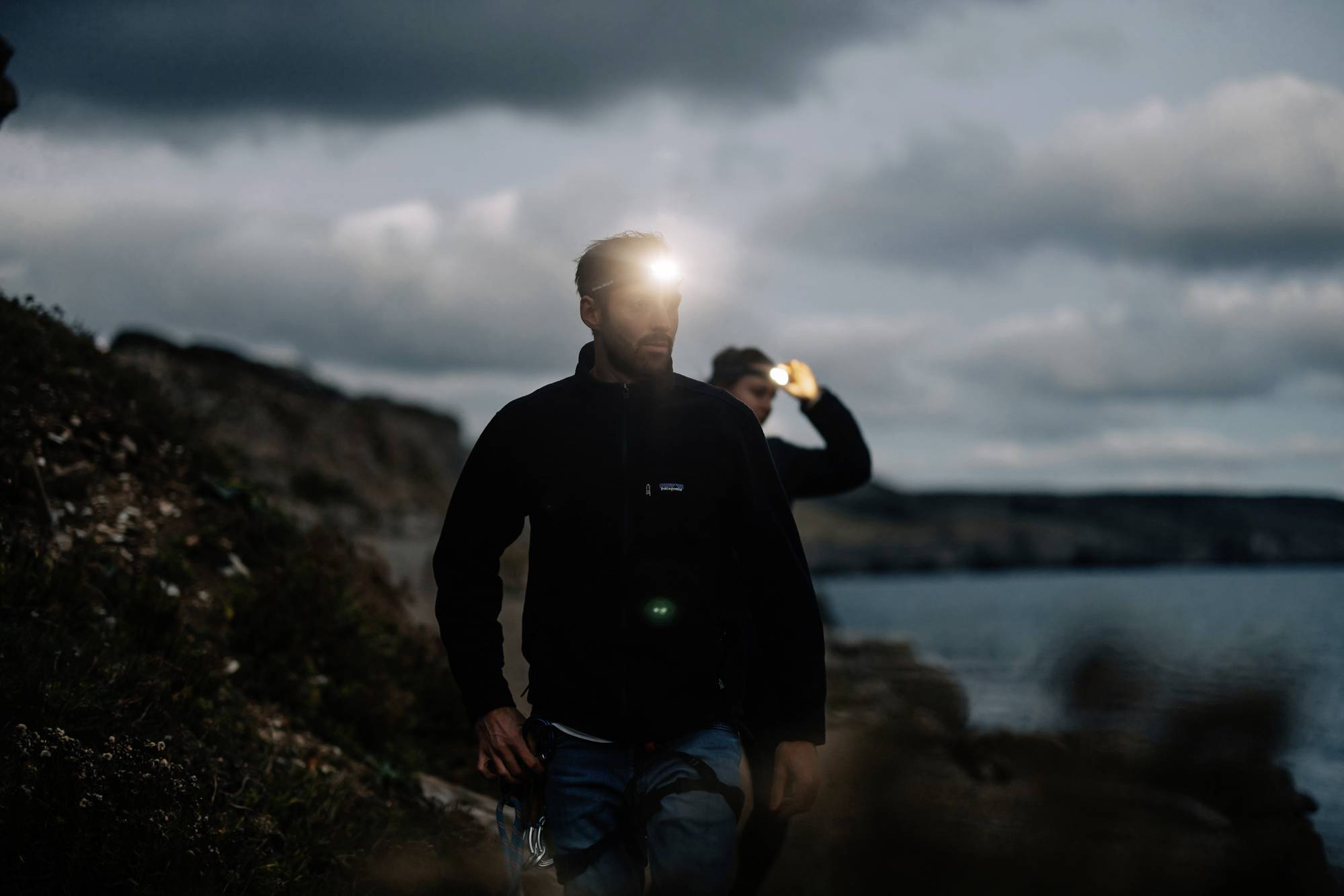 Two people are walking along a coastline in the dark. They are wearing head torches that illuminate their surroundings. The sky is overcast.