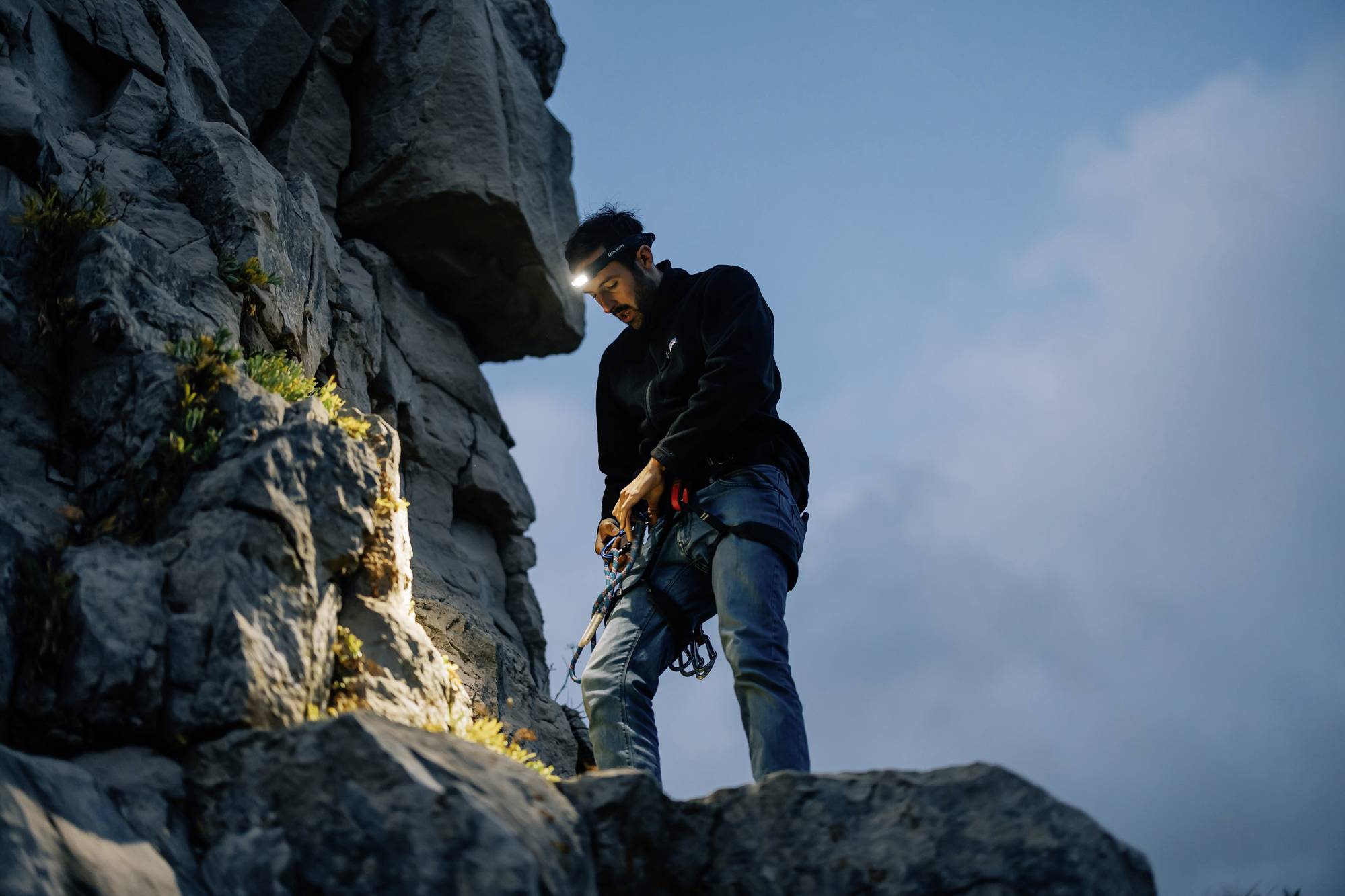 A climber with a headtorch stands on a rock, pulling rope through a belay device. The background shows a cloudy sky at dusk.