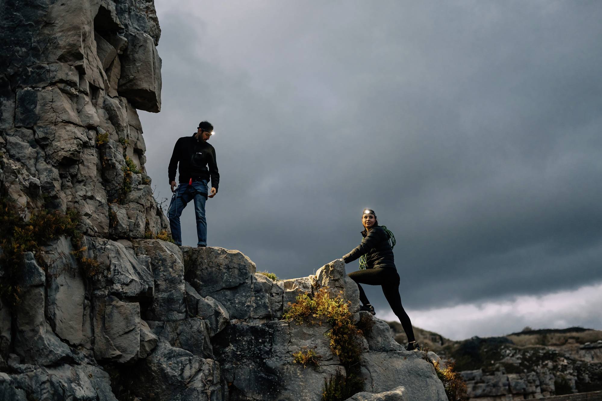 Two people climb a rocky cliff under a cloudy sky. The scene depicts a sense of adventure and physical challenge in nature.