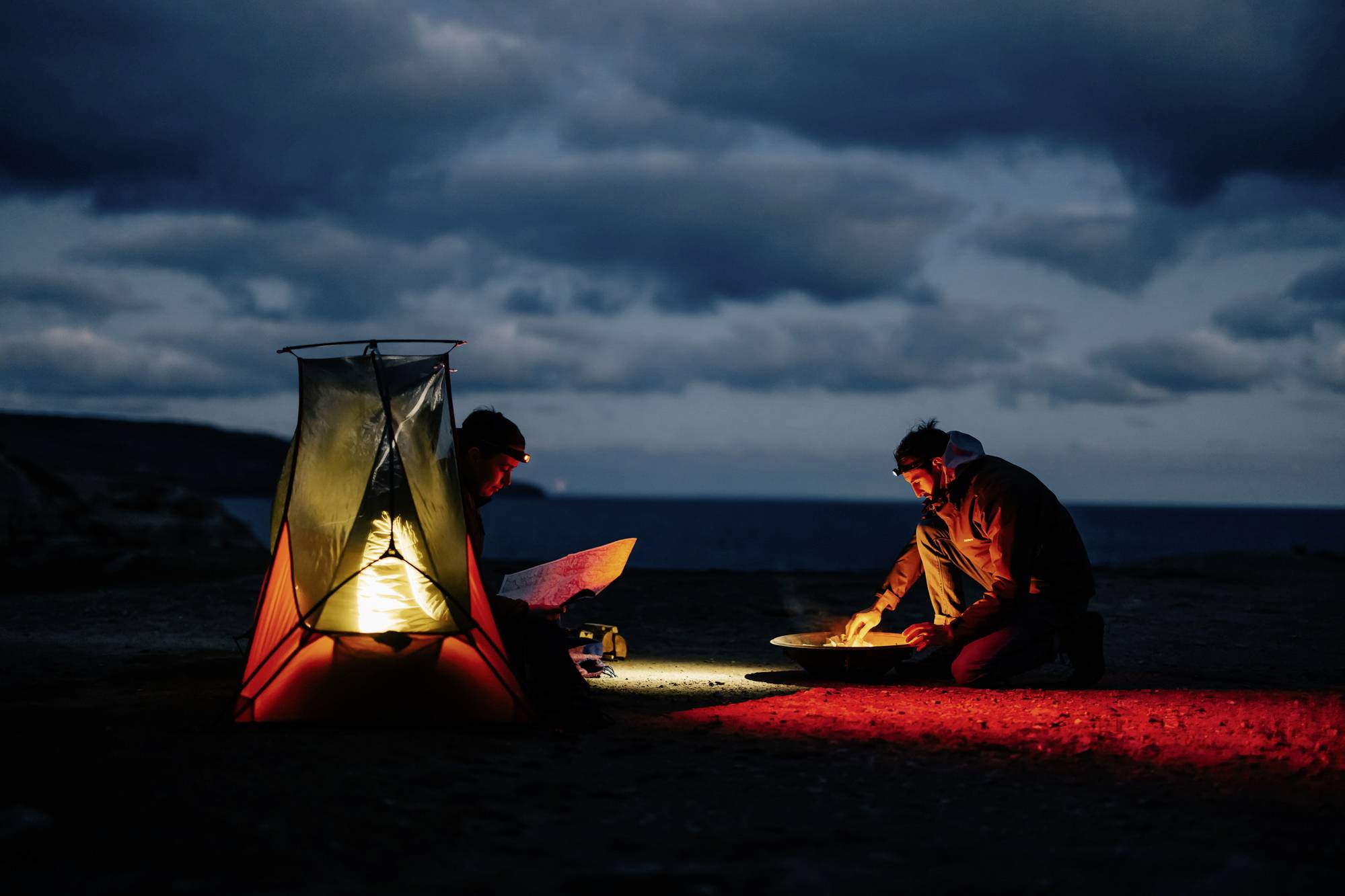 Two people are camping at night by the sea, illuminated by a lantern. One is reading a map, the other is cooking on a portable stove. A cloudy sky is in the background.