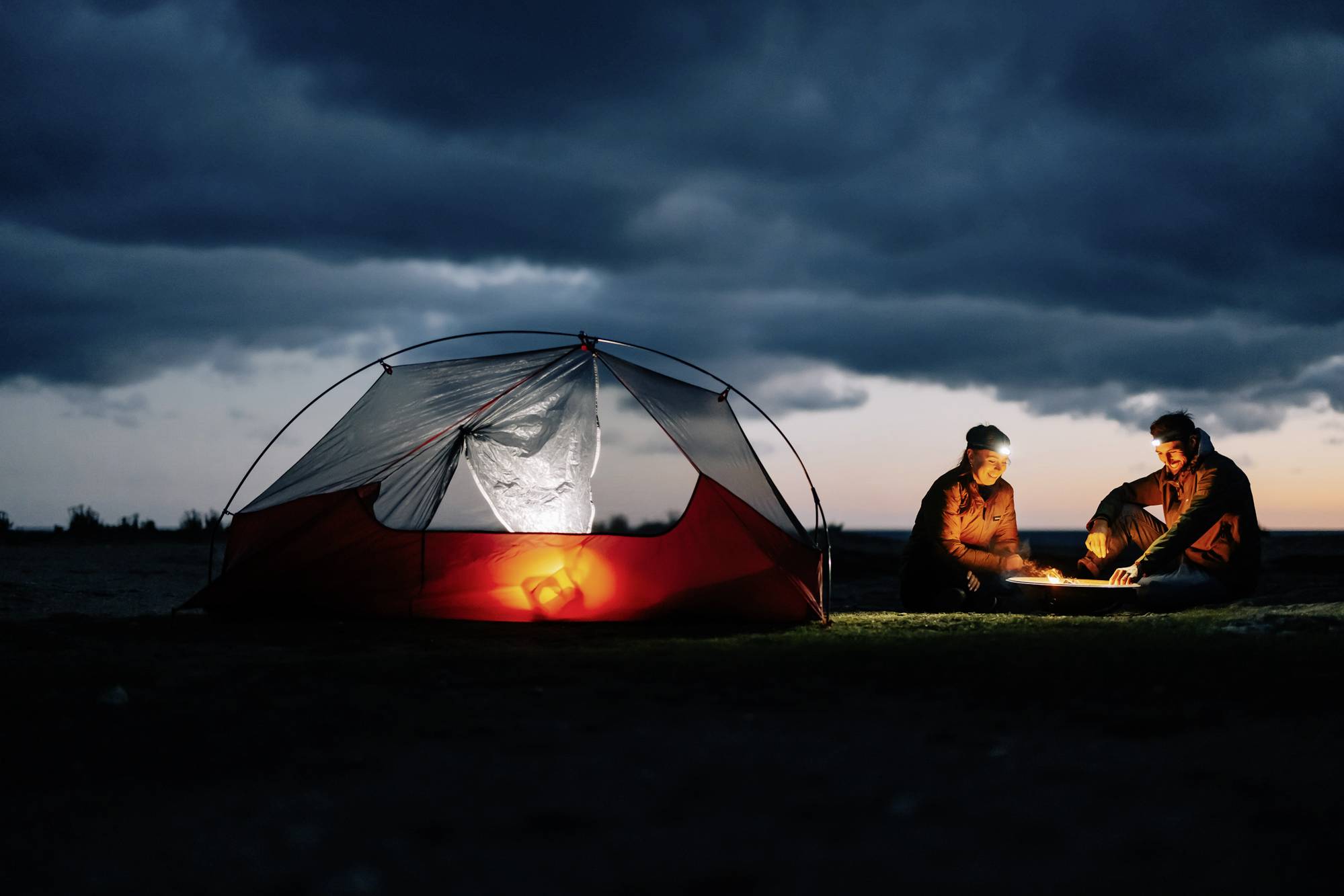 Two people are sitting by a campfire in the evening next to an illuminated tent. Dark sky and tranquil landscape in the background.