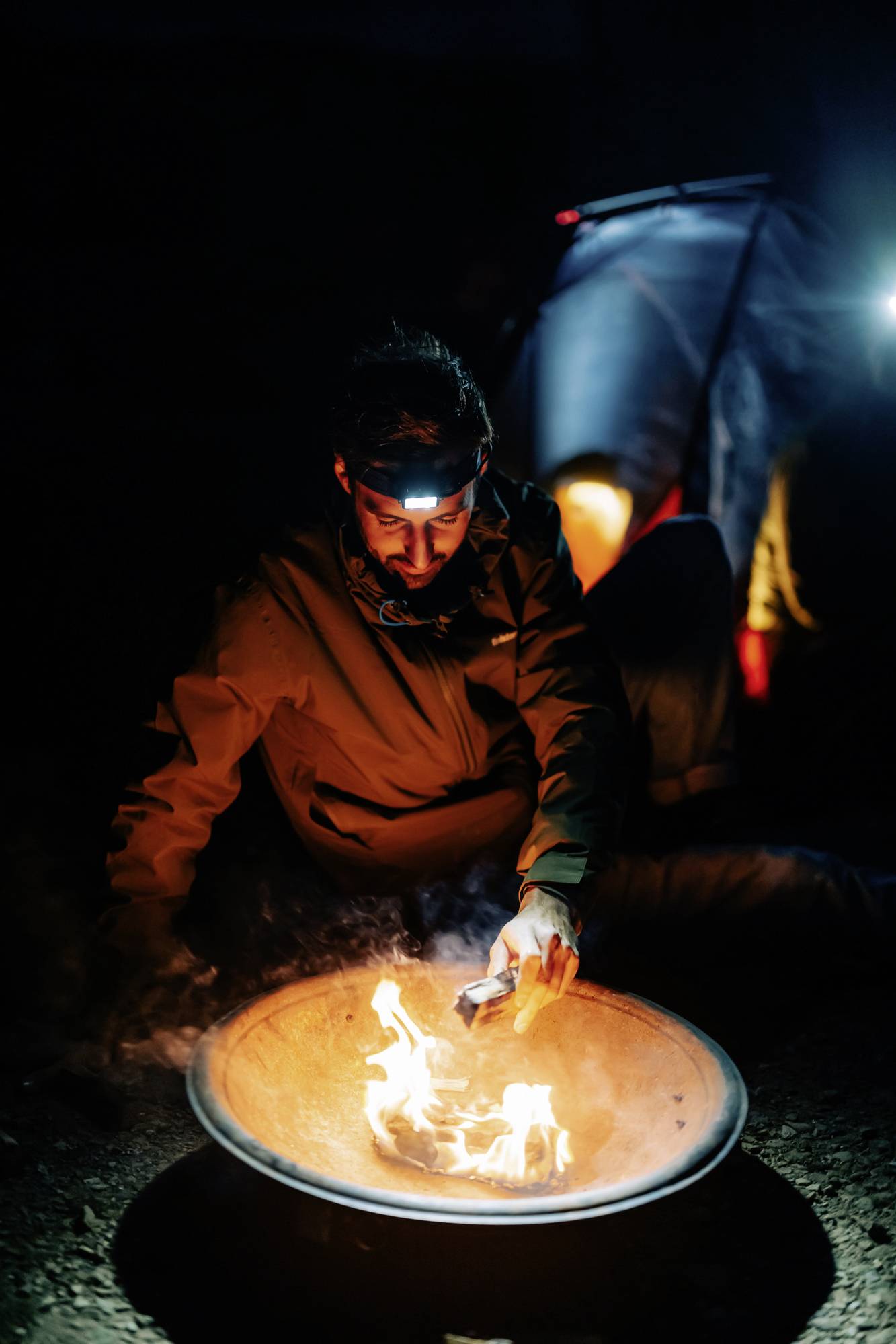 A person sits by a burning fire bowl at night, wearing a head torch. A tent is illuminated in the background.
