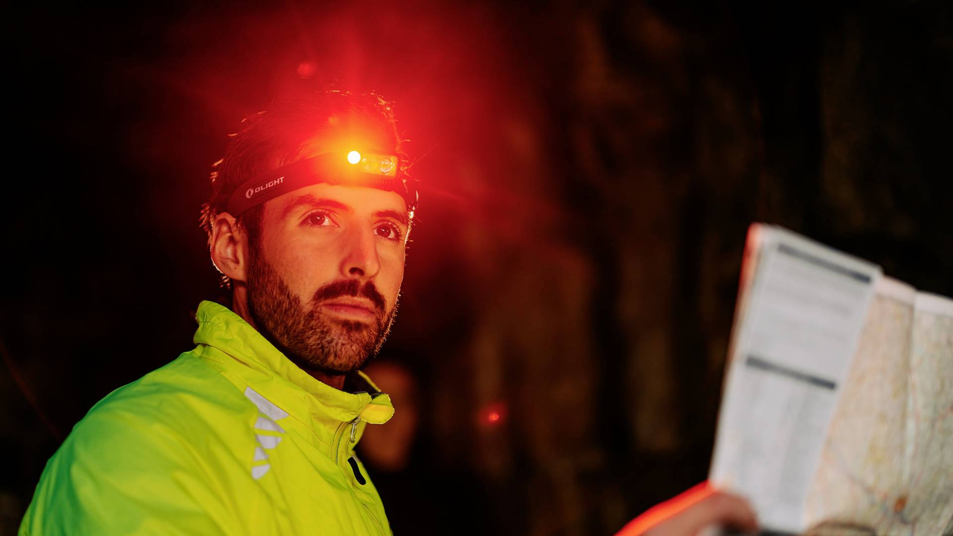 A man wearing a head torch holds a map in his hand. He is wearing a high-visibility, reflective jacket, surrounded by a dark environment.
