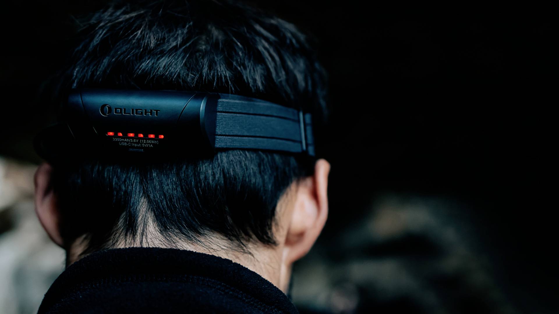 A person with short dark hair is wearing a head torch in the dark. The torch has glowing LED indicators on the back.