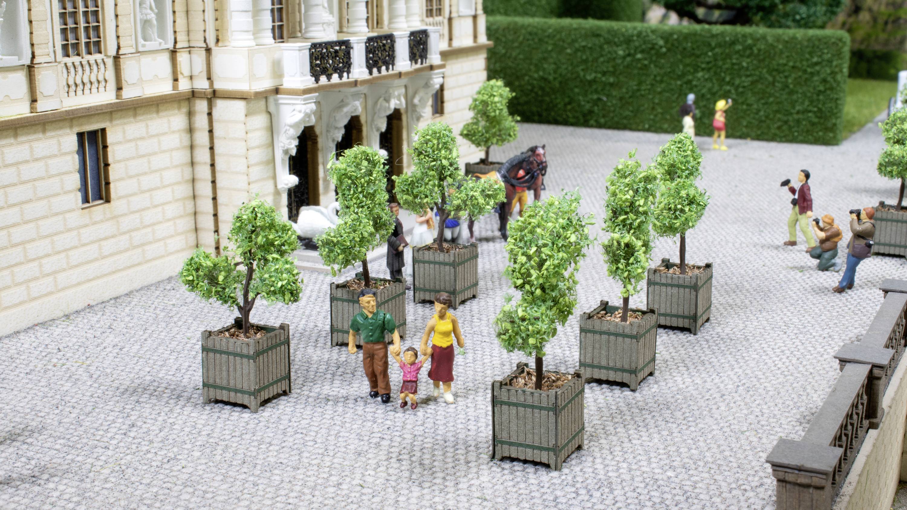 A miniature model shows people walking in a cobbled courtyard between potted trees. A couple are holding the hands of a child.