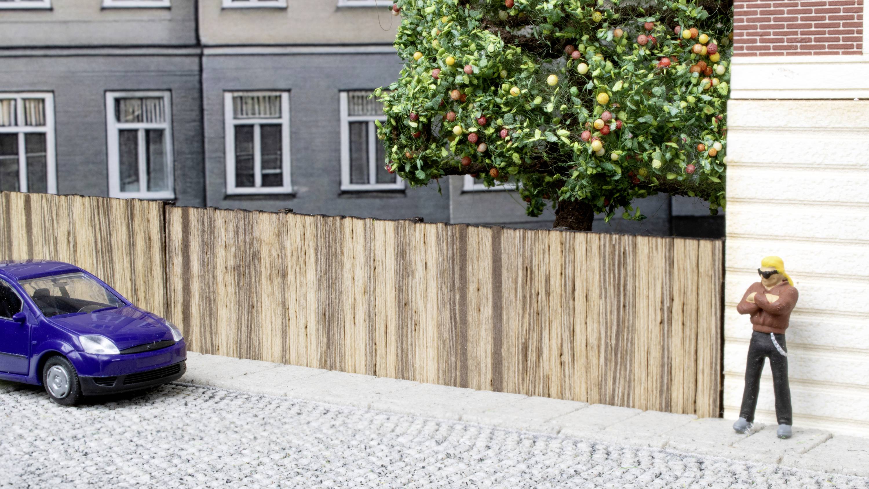 A model of a neighbourhood with a miniature car and figure on a cobblestone street, with a tree beside it.