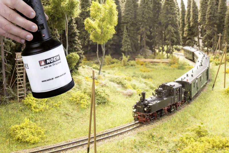 A small model train travels through a lush, forested landscape, with a hand holding a light positioned above for scenery enhancement.